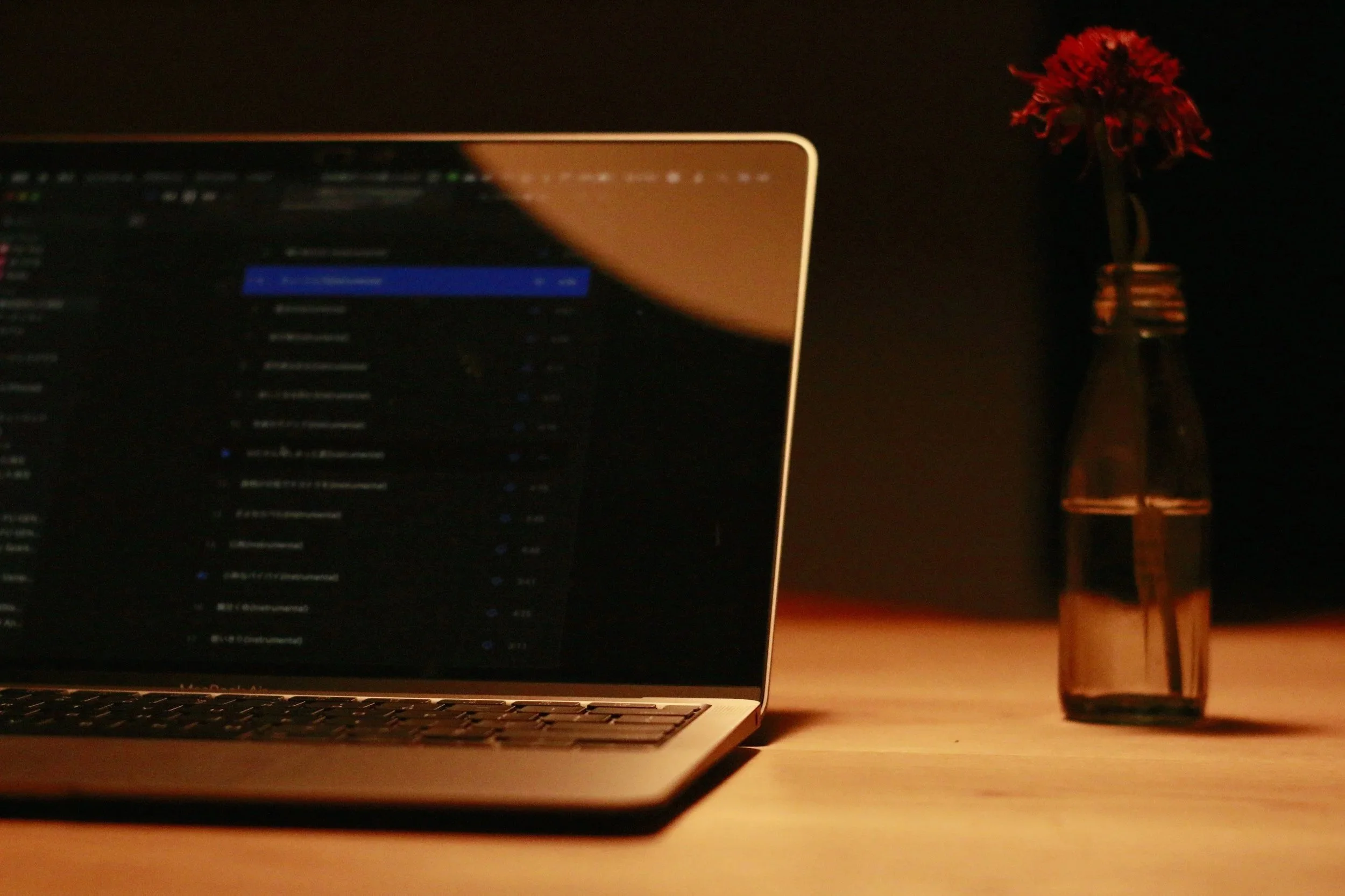 A laptop with an open screen displaying a blurred dark interface, next to a brown glass bottle with a red flower inside, on a wooden surface in a dimly lit setting.