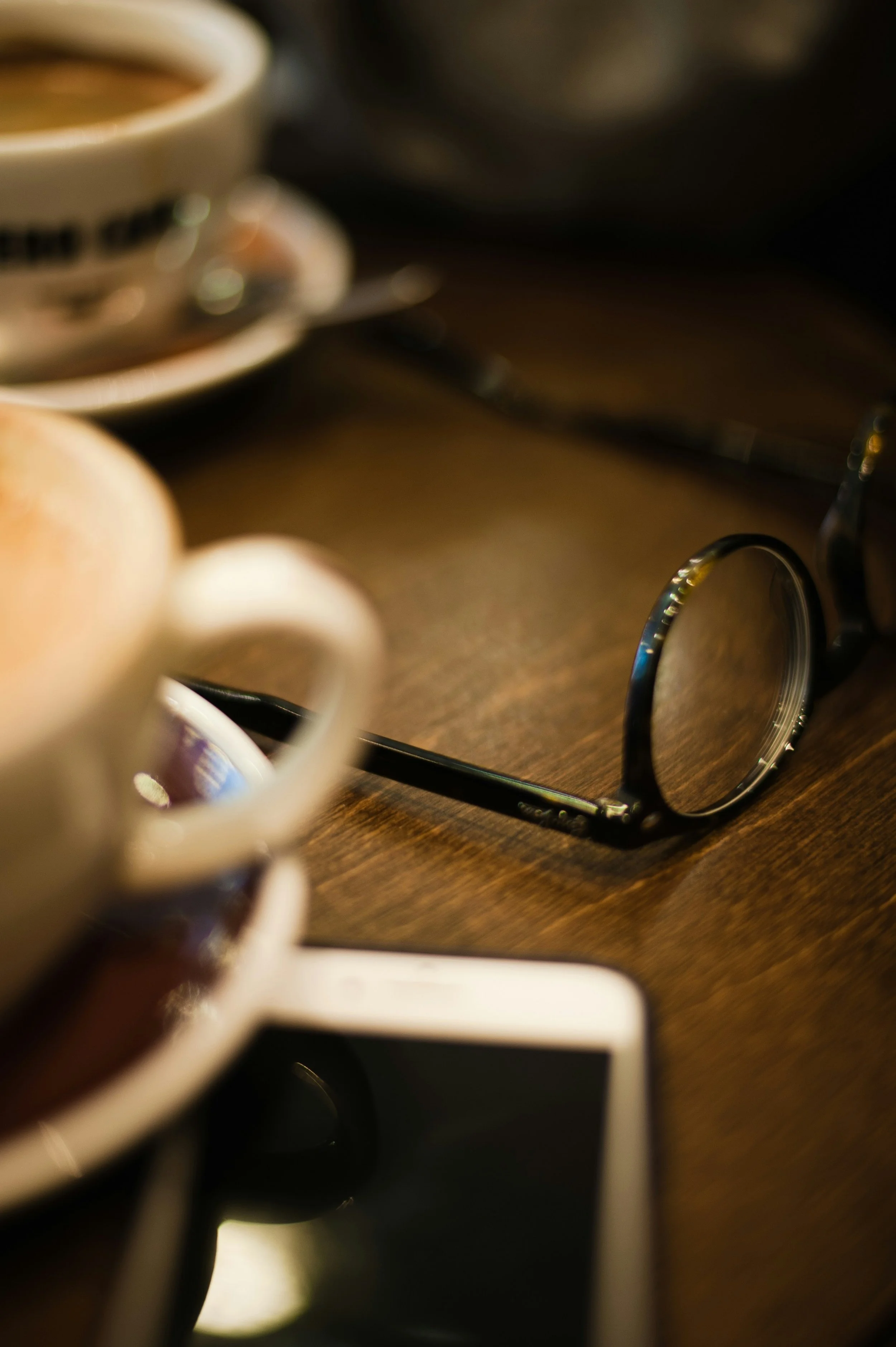 A close-up photo of glasses resting on a wooden surface, with a cup of coffee or tea and a blurred spoon in the background.