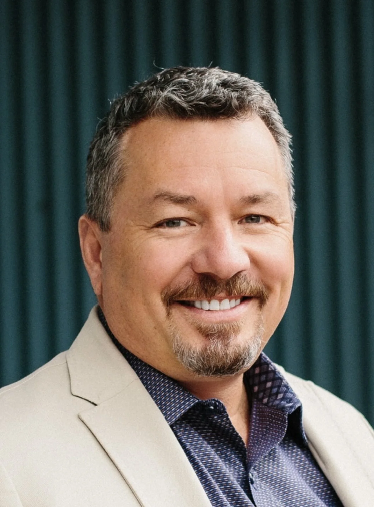 A smiling man with short, dark, and gray hair and facial hair, wearing a light-colored blazer and a dark patterned shirt, against a dark teal curtain background.