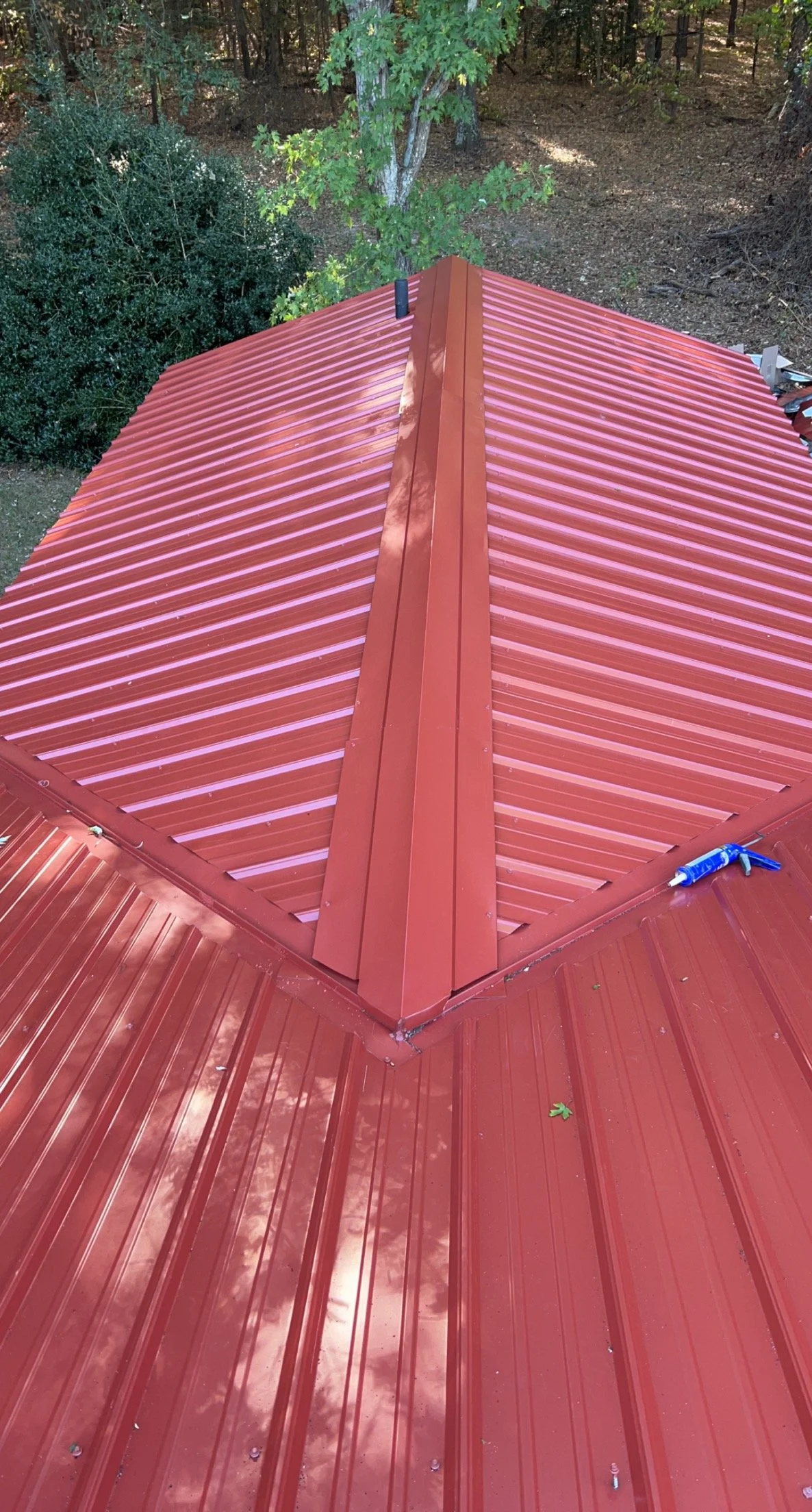 Red metal roof with a central ridge and multiple panels, surrounded by trees and greenery.