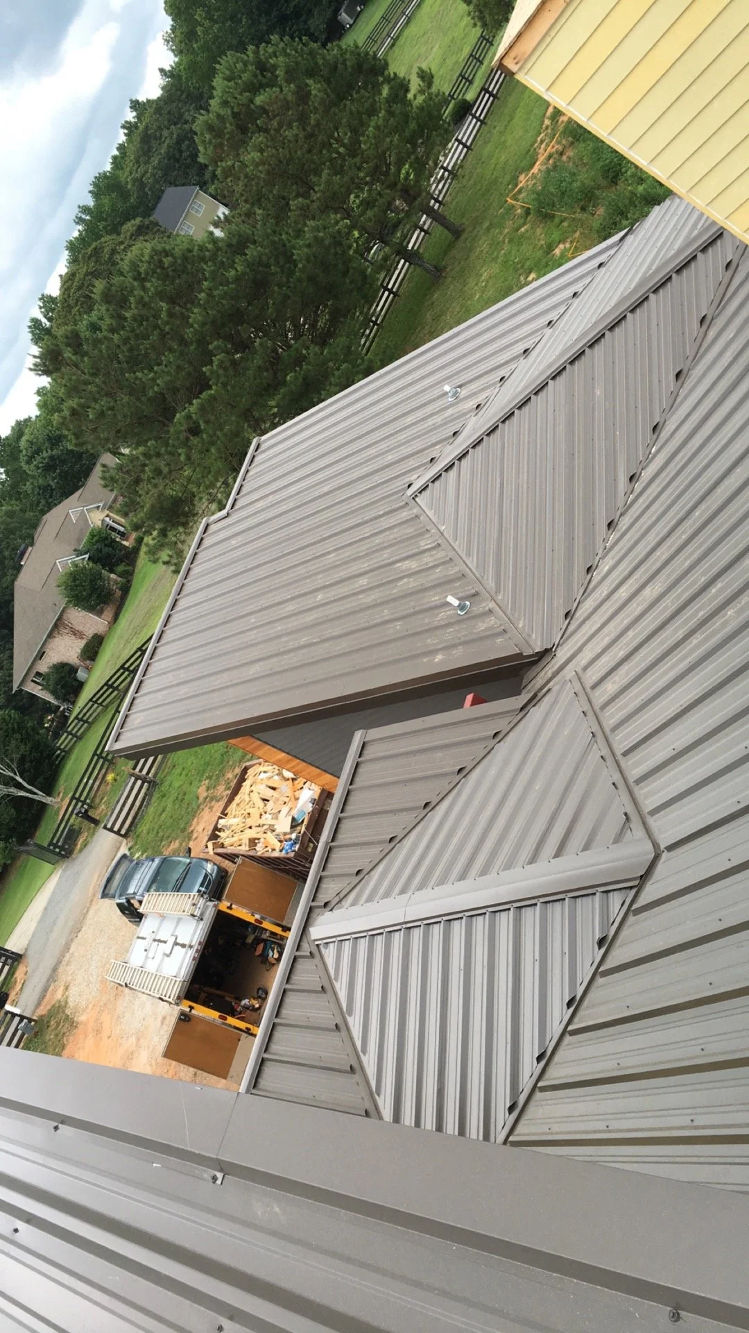 A view of metal roofs on a residential building, with trees, a fence, a yard, and a truck with construction materials in the background.