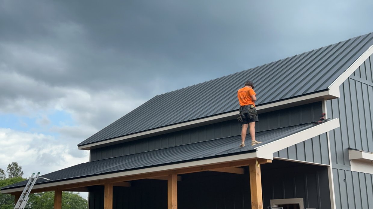 Construction worker in orange shirt installing a metal roof on a house with dark roofing panels and wooden support beams.