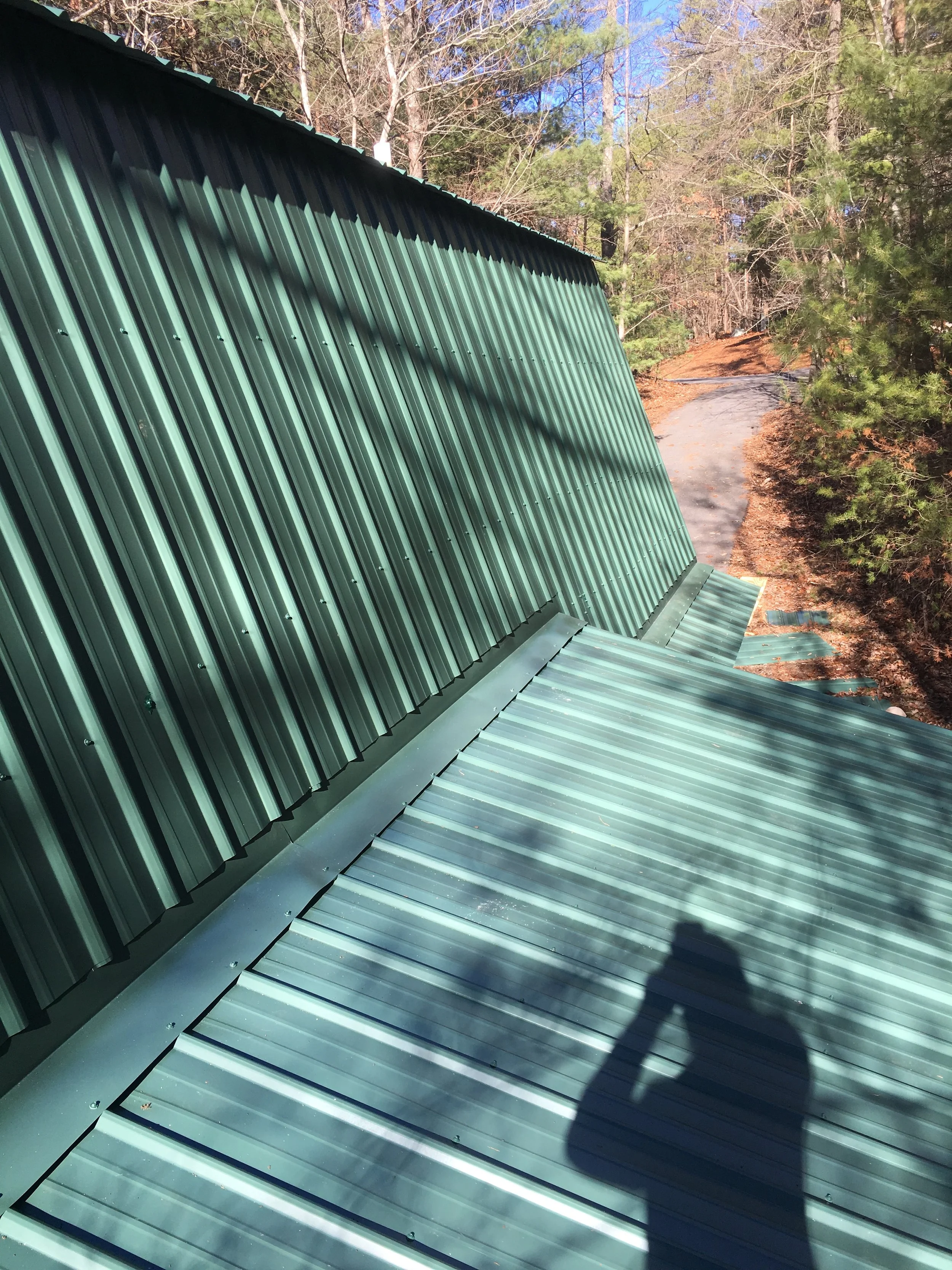 Close-up of a green metal roof with ridges, with trees and parking lot in the background, and the shadow of the person taking the photo visible on the roof.
