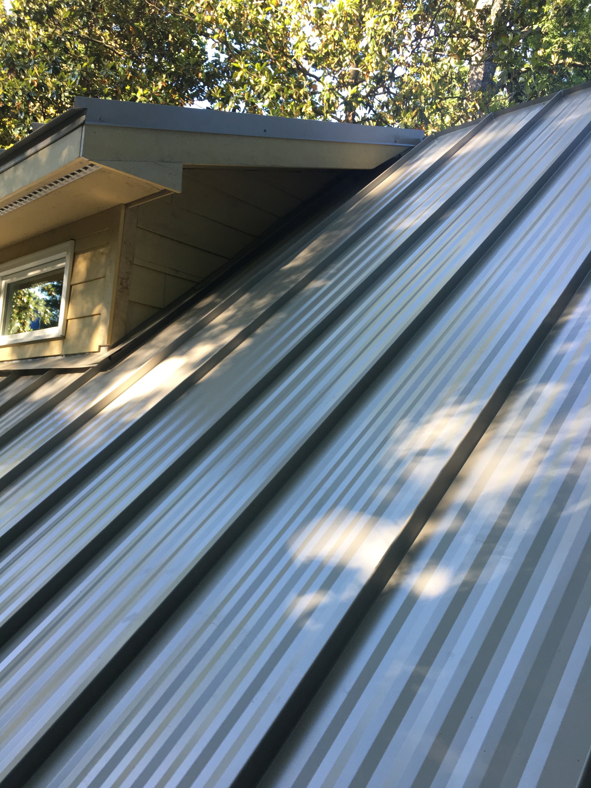Close-up of a metal roof with vertical panels on a house, with trees and sky in the background.