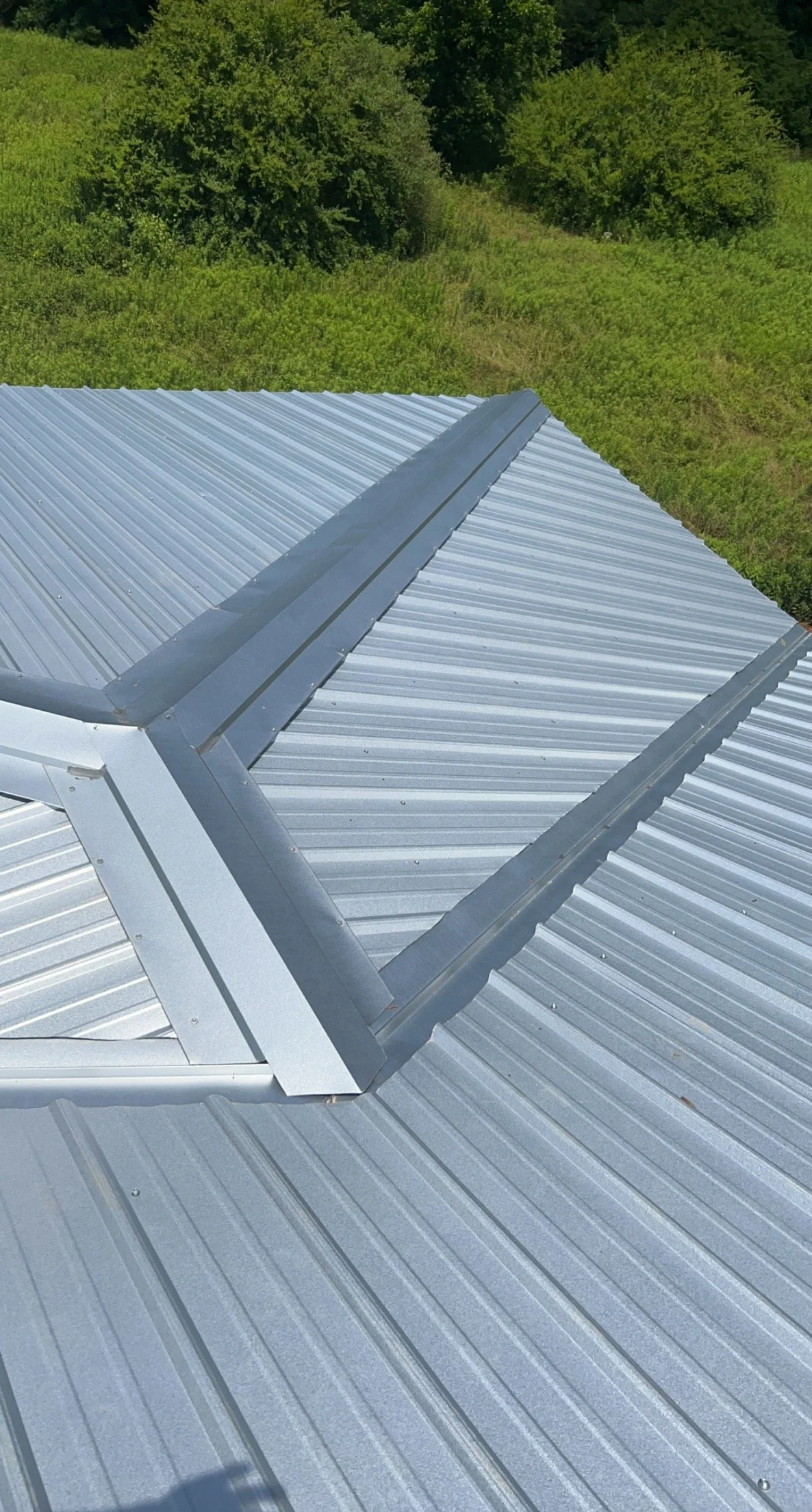 Close-up of a metal roof with a ridge vent, surrounded by green trees and grass.