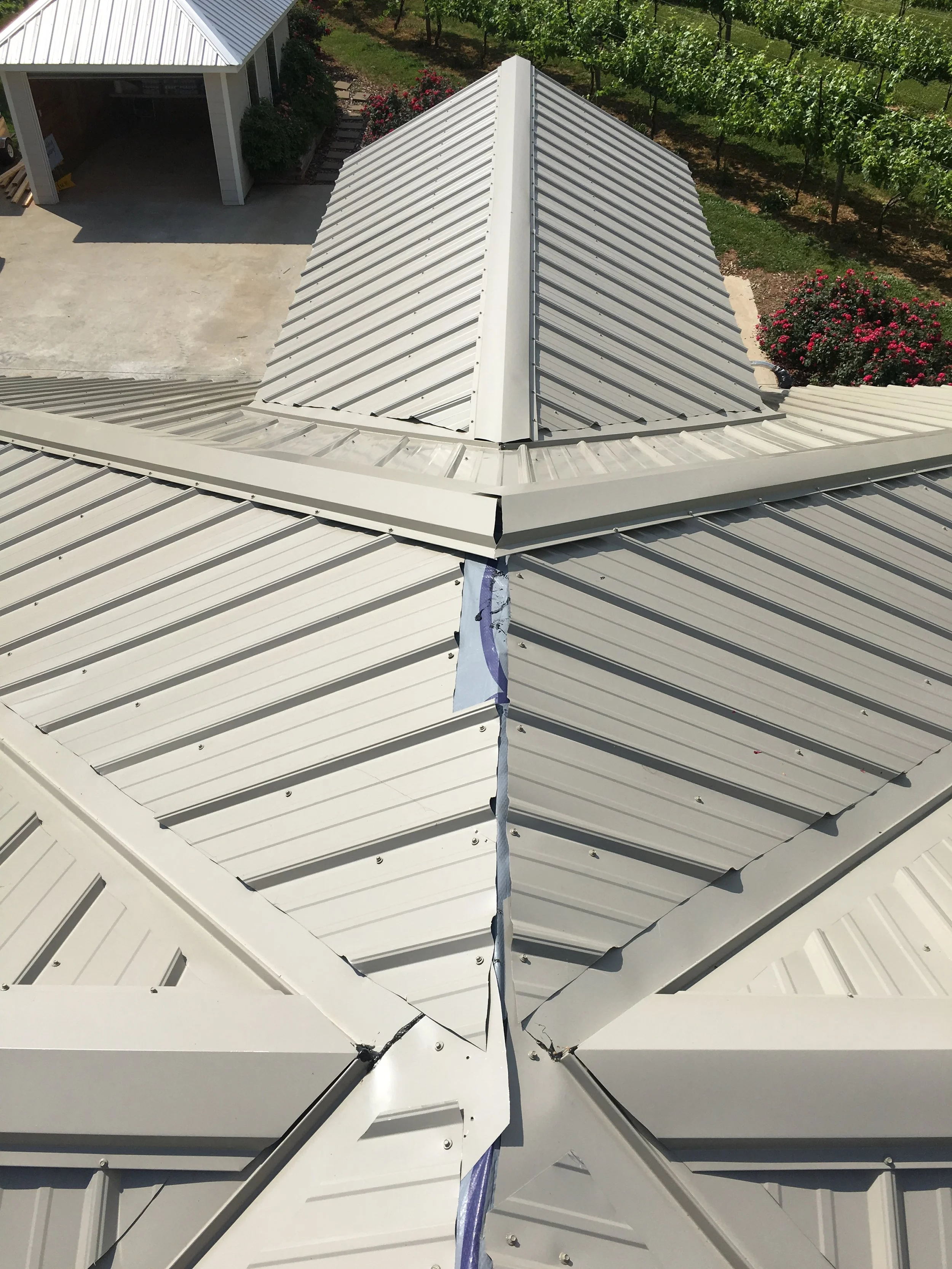 A photograph of a metal building roof with multiple sections, featuring a metal peak at the center and ongoing construction or repair work with a strip of cloth hanging from the seam.