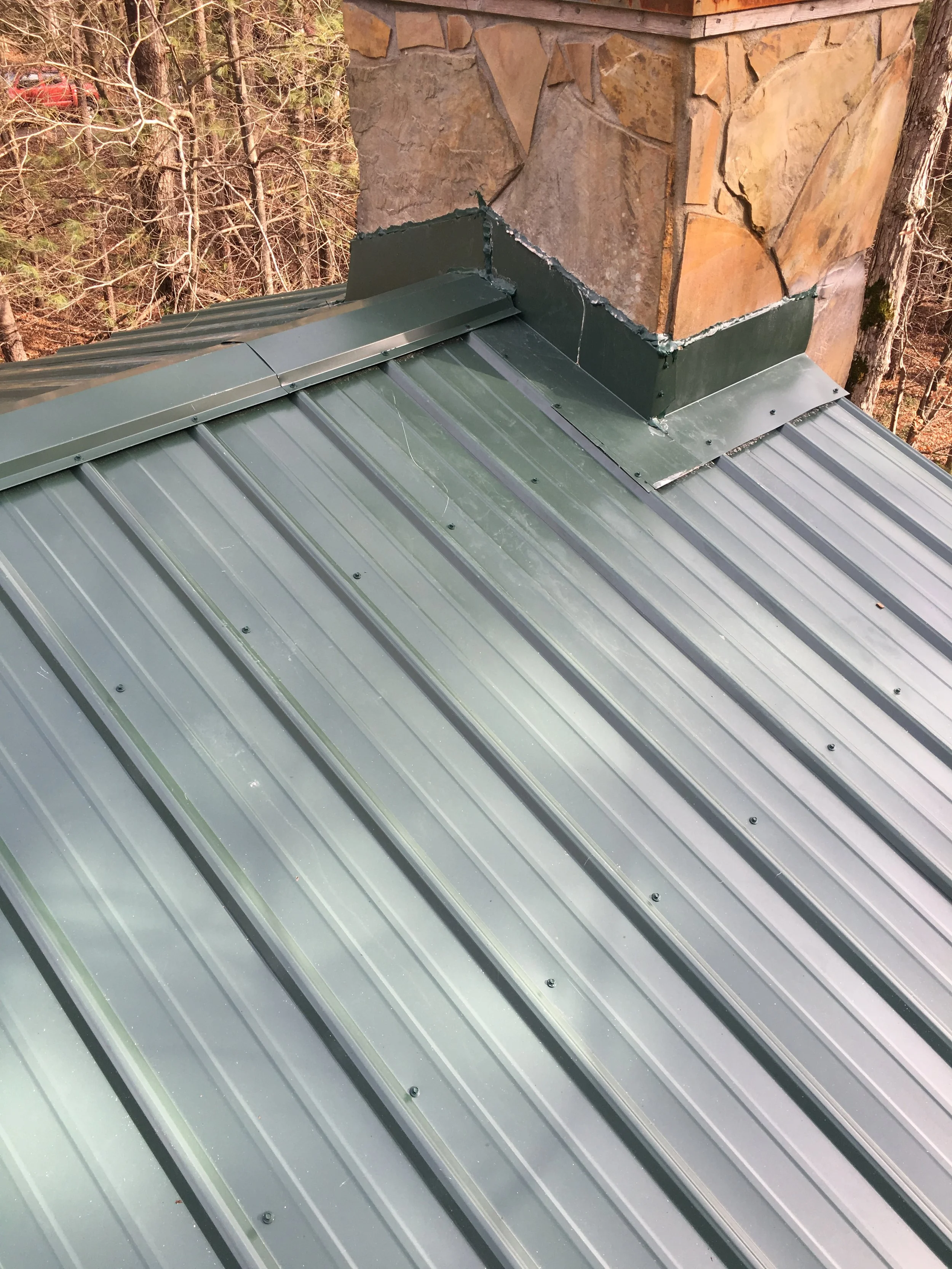 Close-up of a green metal roof with a chimney and stone exterior in the background, surrounded by trees.