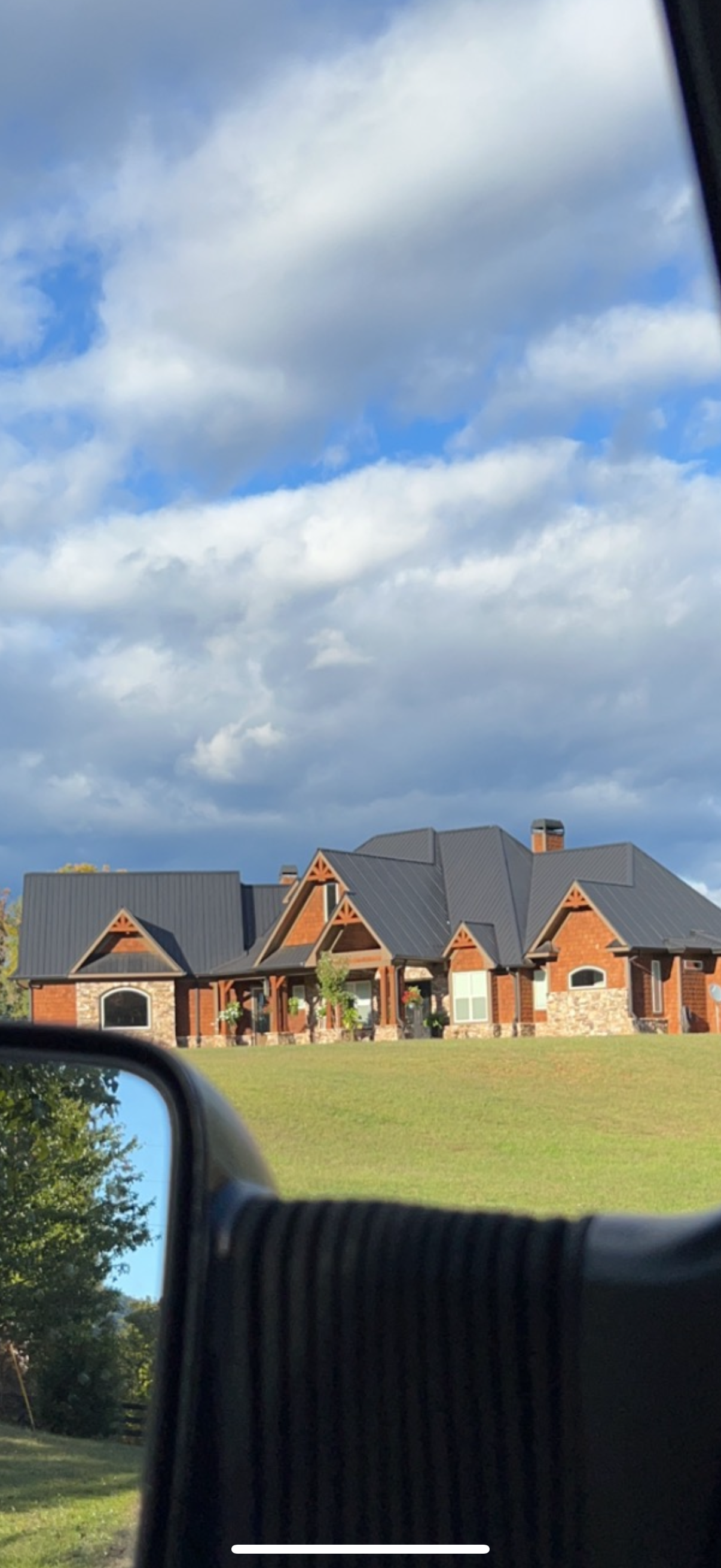 View of a large brick house with a dark metal roof, situated on a grassy lawn under a partly cloudy sky, photo taken from inside a vehicle.