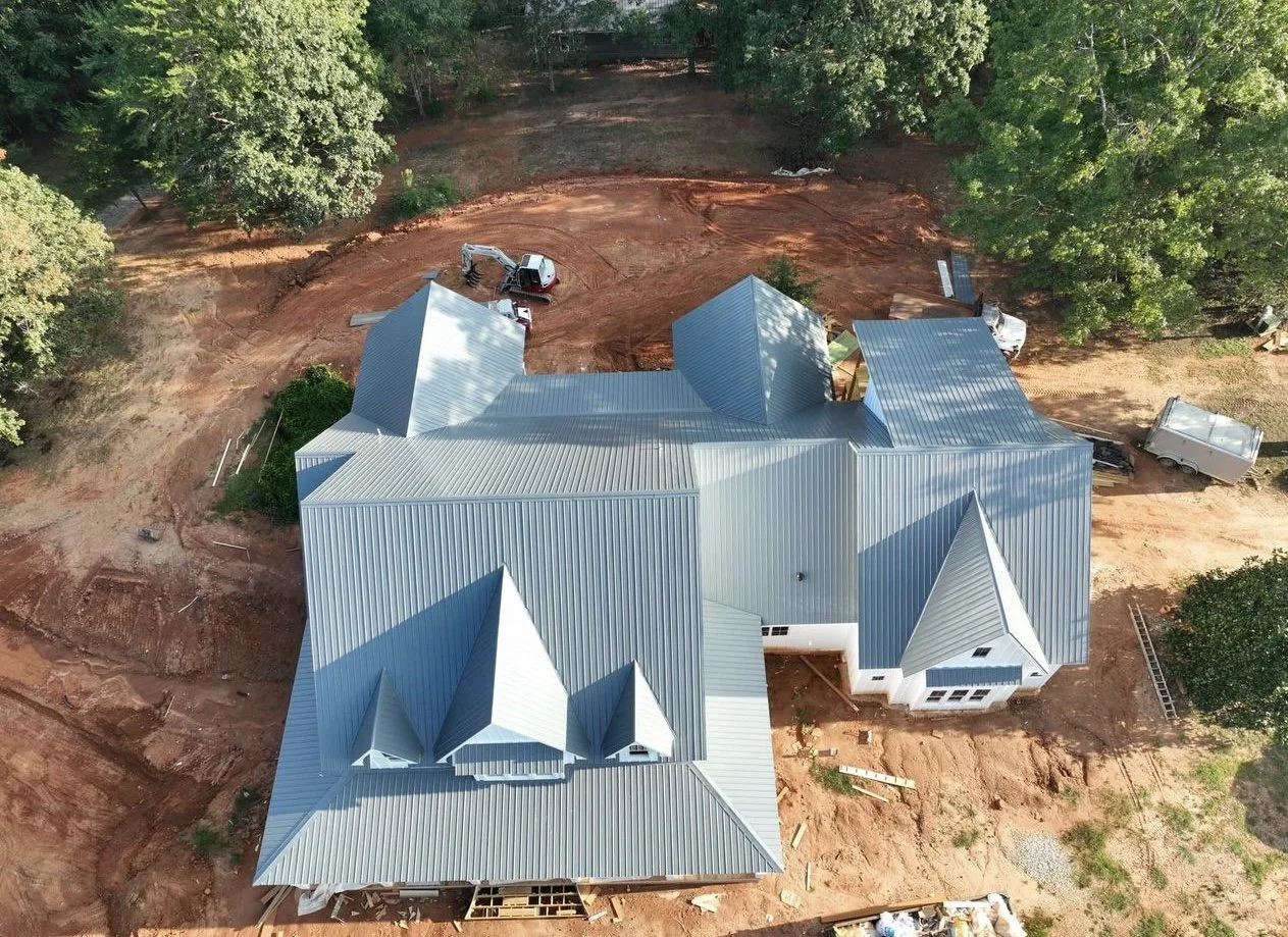 Aerial view of a large house under construction with a metal roof, surrounded by trees and construction equipment on bare ground.