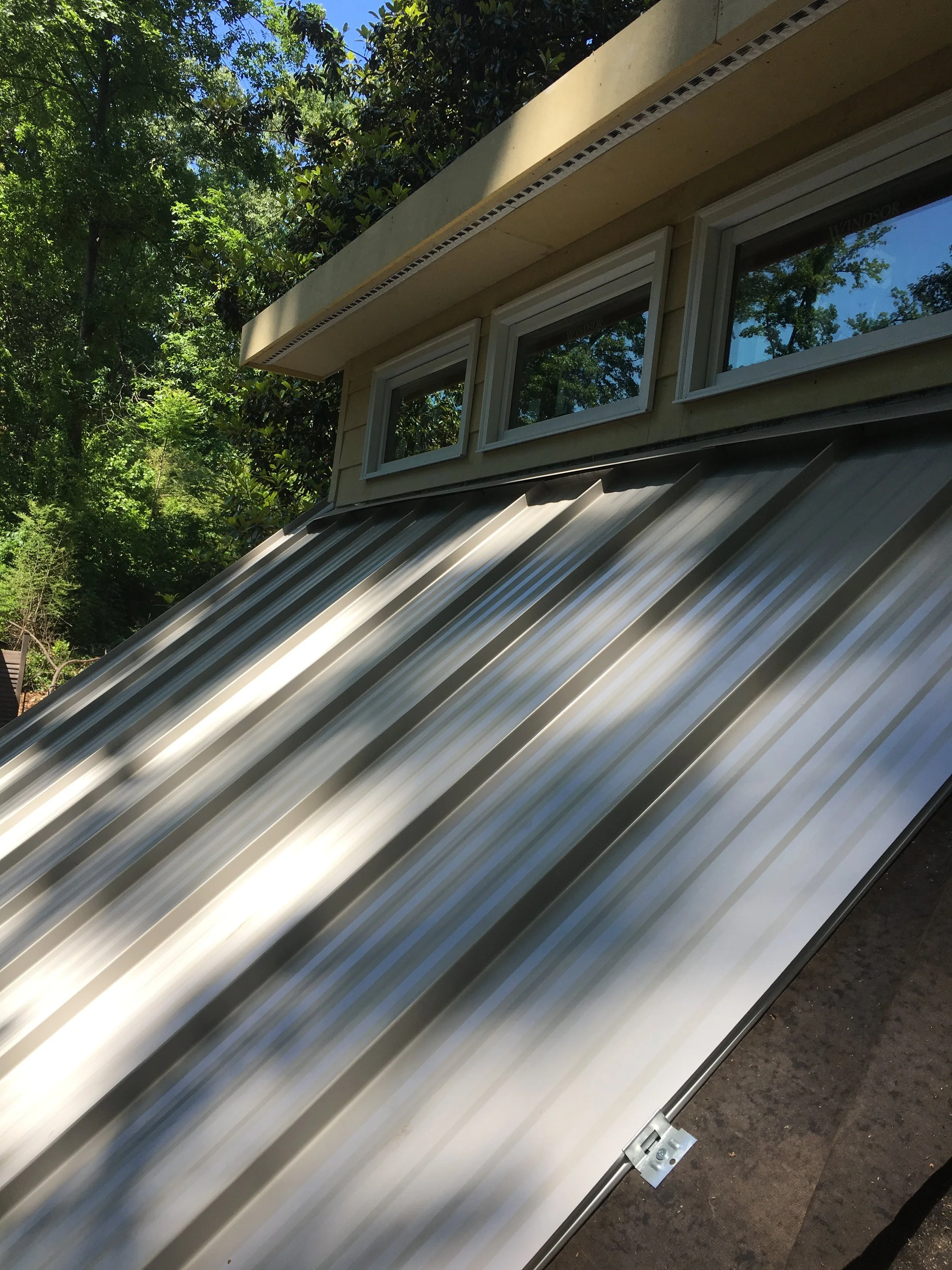 Metal roof with three small windows above, part of a beige building surrounded by green trees on a sunny day.
