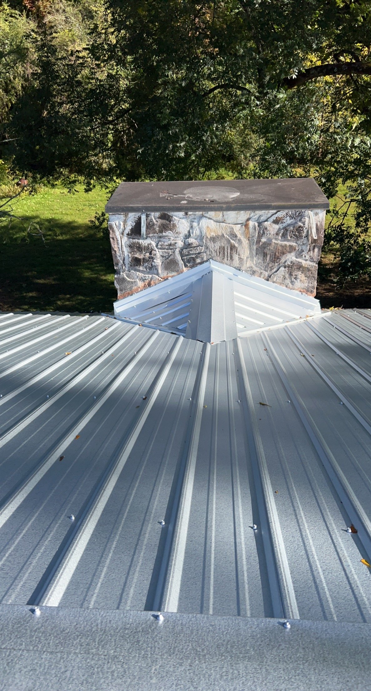 Metal roof with a central vent flashing and a stone chimney in the background, surrounded by trees.