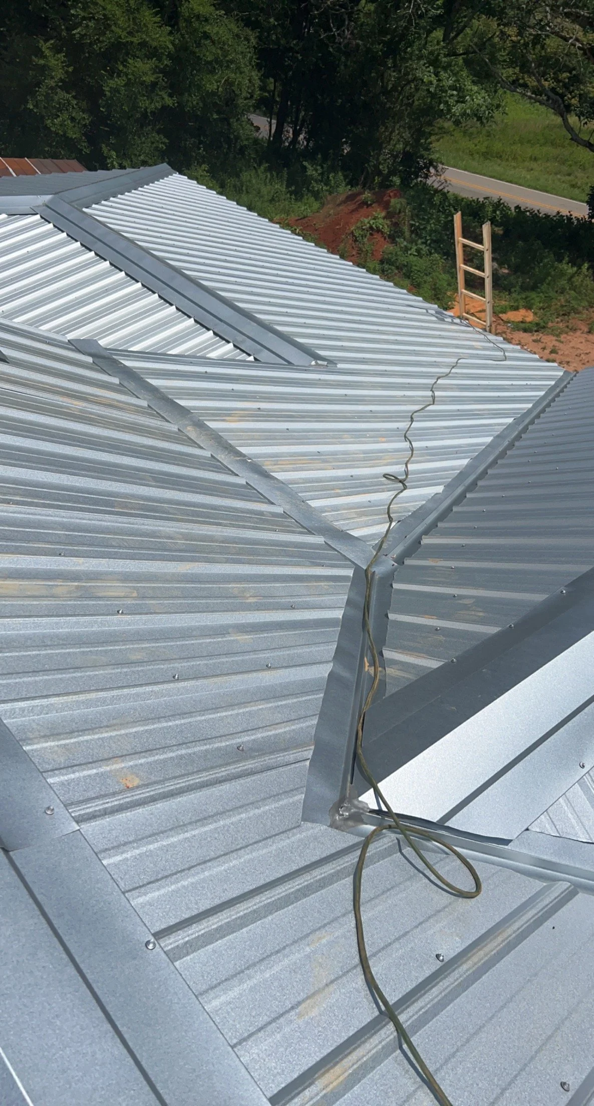 Metal roof panels with a crack in the flashing and an electrical cord on top, surrounded by green trees and a road in the background.