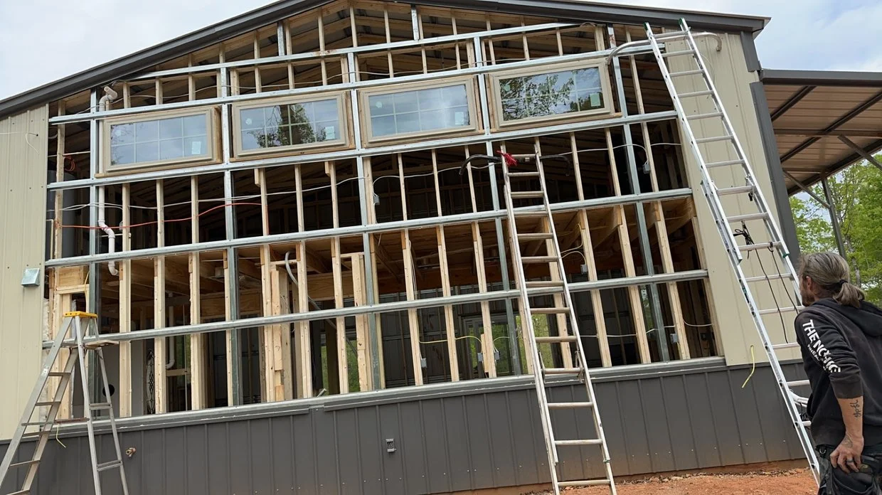 Construction workers are building the frame of a new second-story wall on a house with metal ladders on both sides, and a woman with tattoos observing.