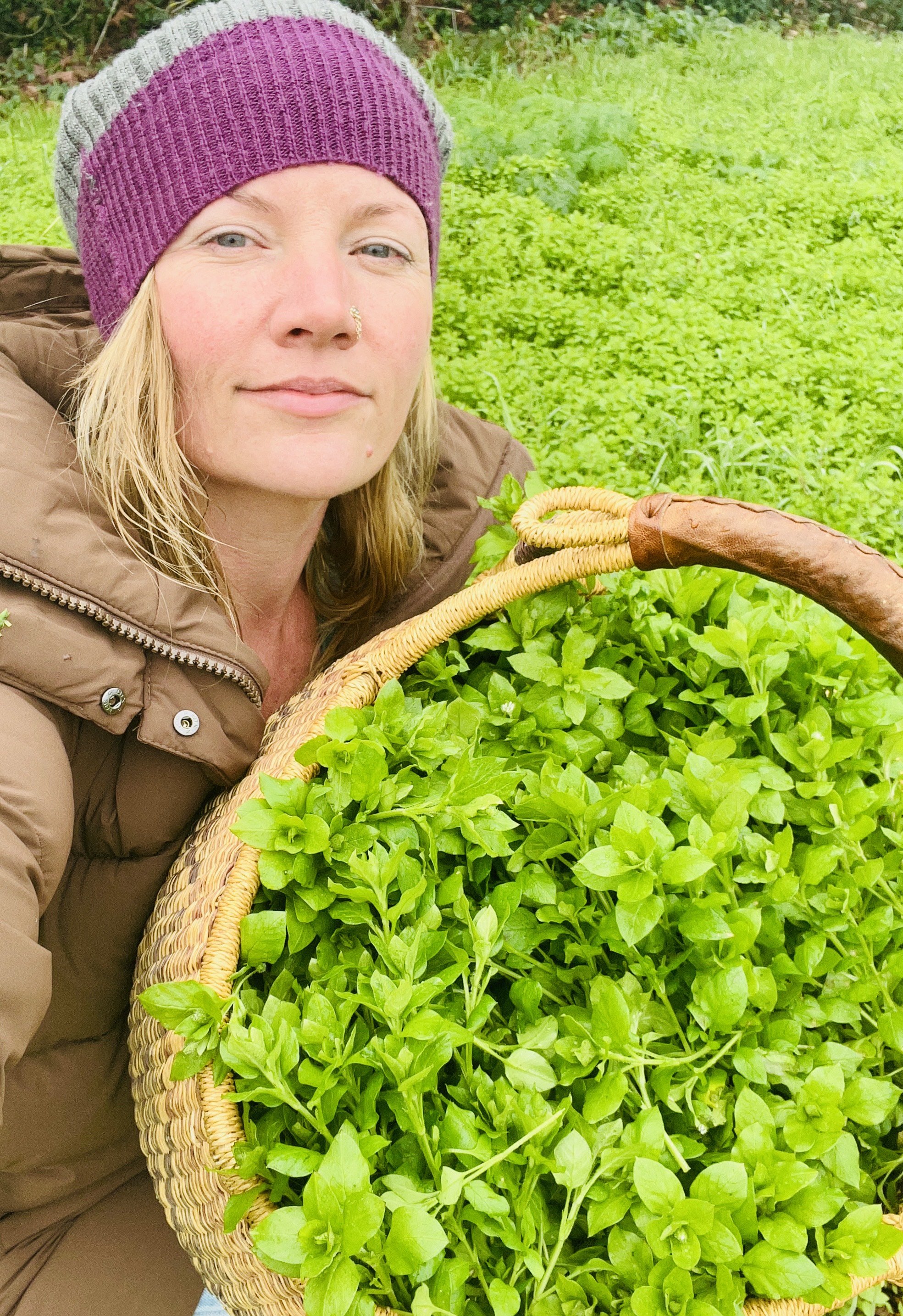 A woman with blonde hair wearing a pink and gray knit beanie and a tan jacket, holding a basket full of fresh green herbs, in a lush outdoor garden.