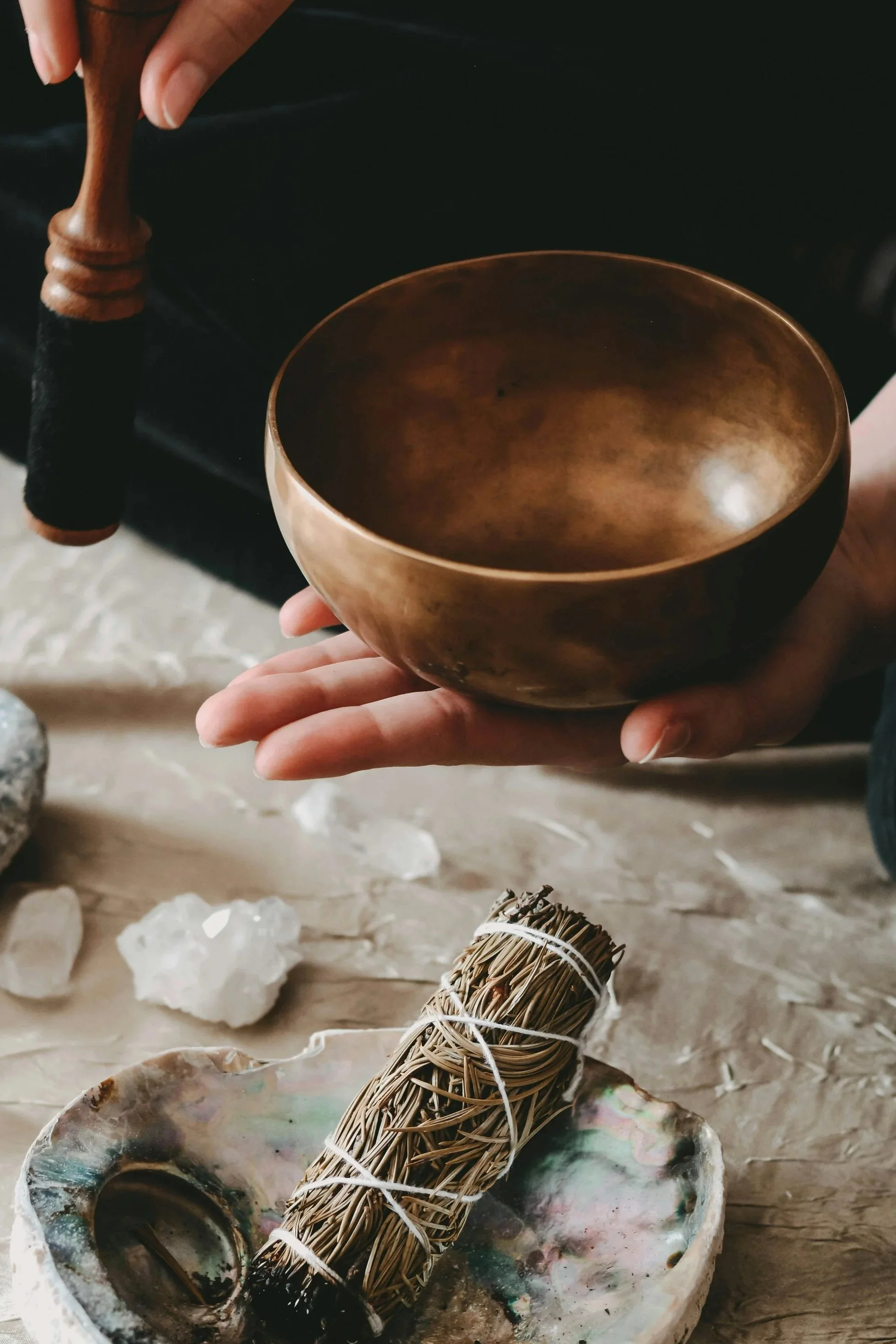 Person holding a singing bowl above a table with a smudge stick, crystals, and a shell dish.