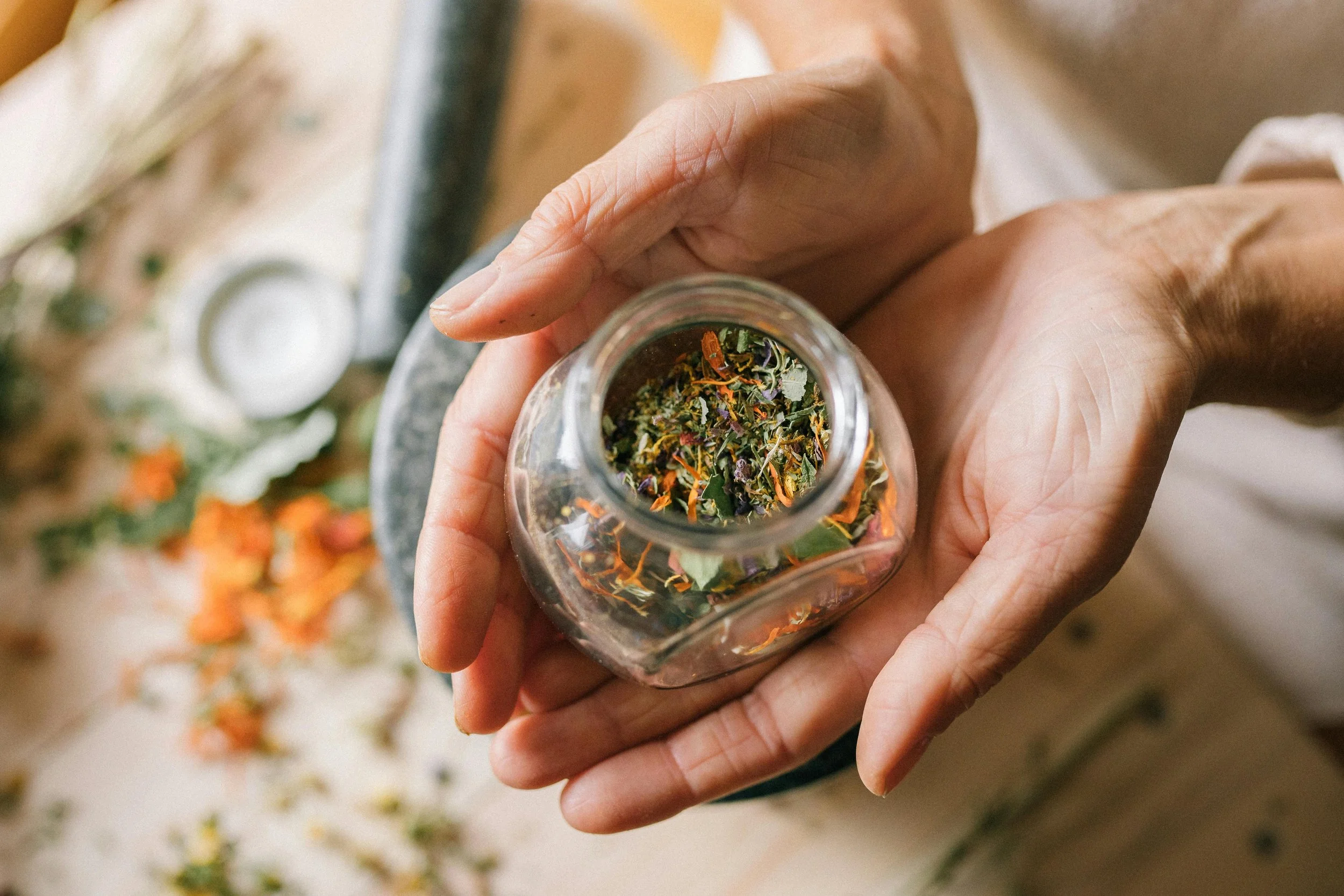 A person holding a glass jar filled with dried herbal flowers and leaves, with some herbs and flowers scattered on a surface below.