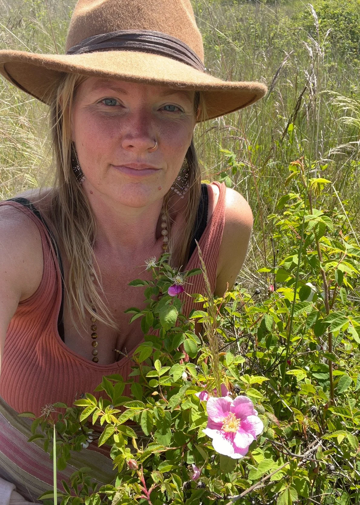 A woman in a wide-brimmed hat and sleeveless top crouches in a field of tall grass and flowering bushes, taking a selfie.