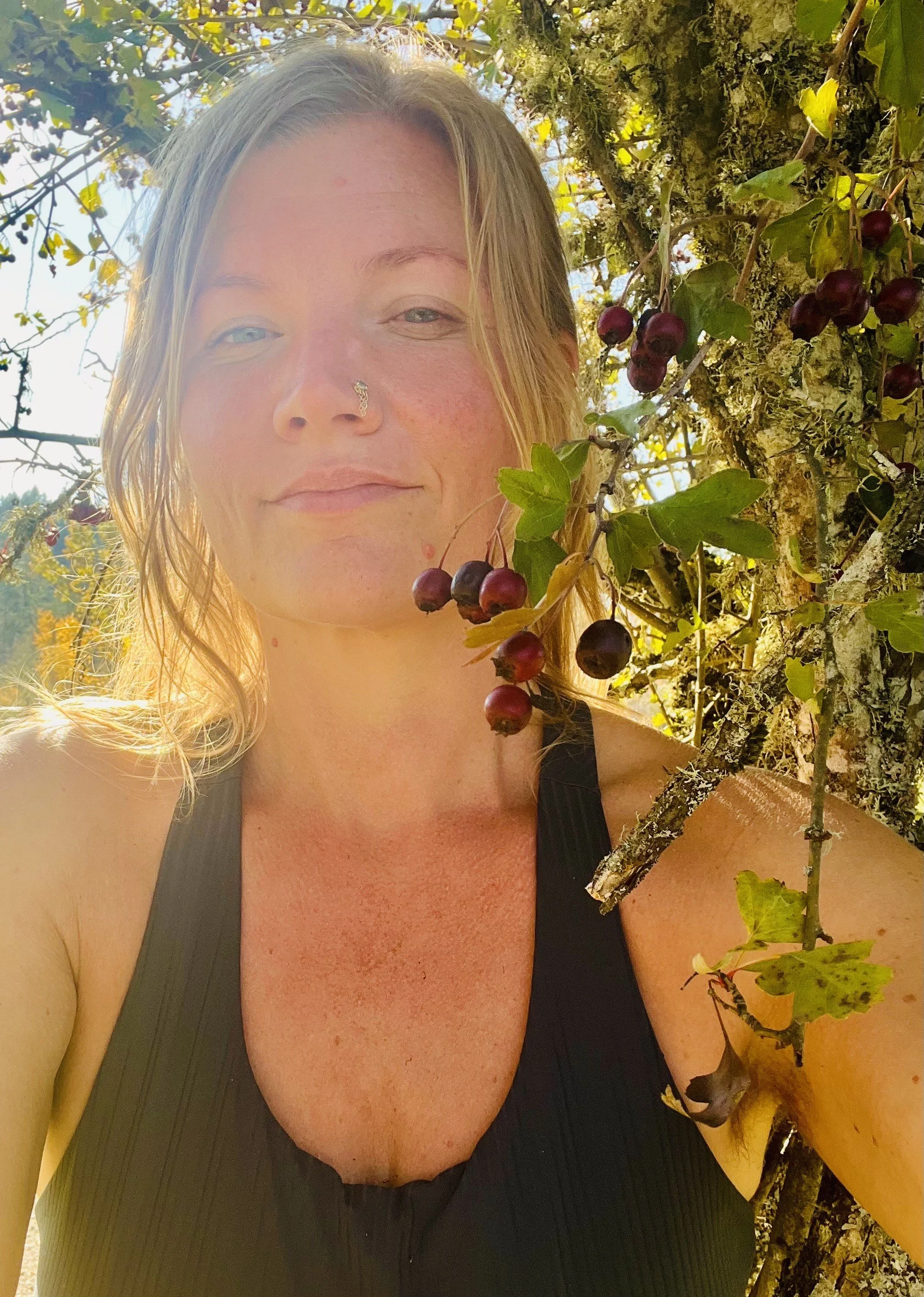 A woman with blonde hair, wearing a black tank top, standing next to a vine with purple berries, outdoors with sunlight.