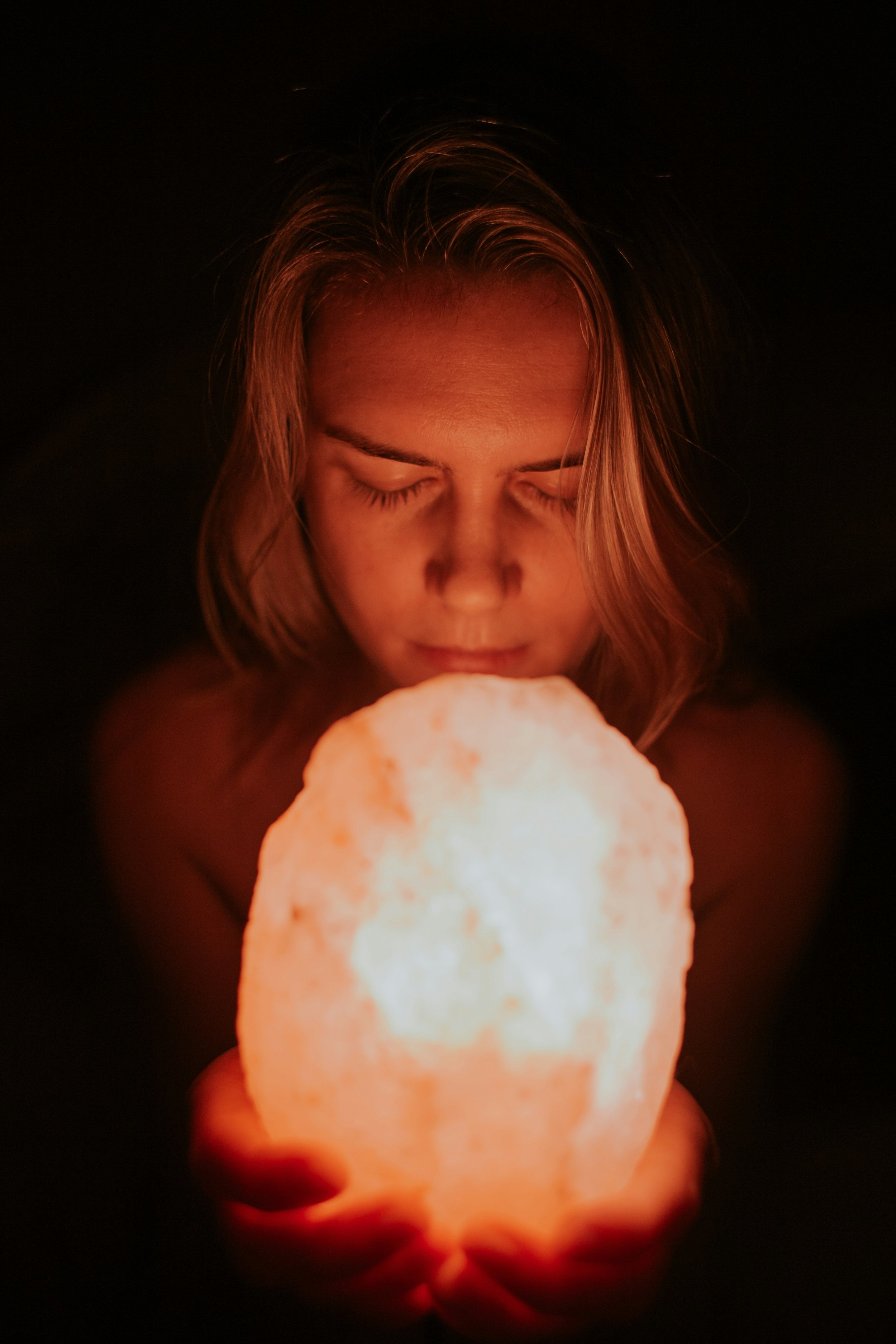 A young woman with closed eyes holding a glowing salt lamp in a dark room.