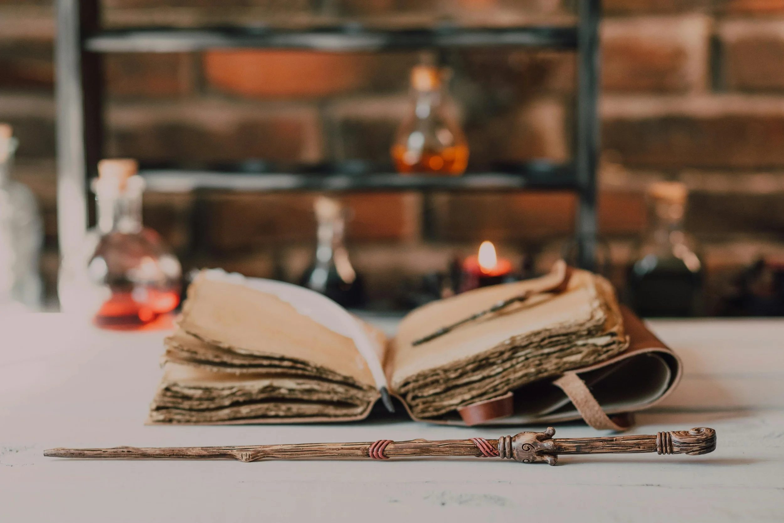 An open old, weathered book lying on a white surface with a rustic wooden staff in front of it. In the background, there are glass bottles with liquids, a small candle, and a brick wall.