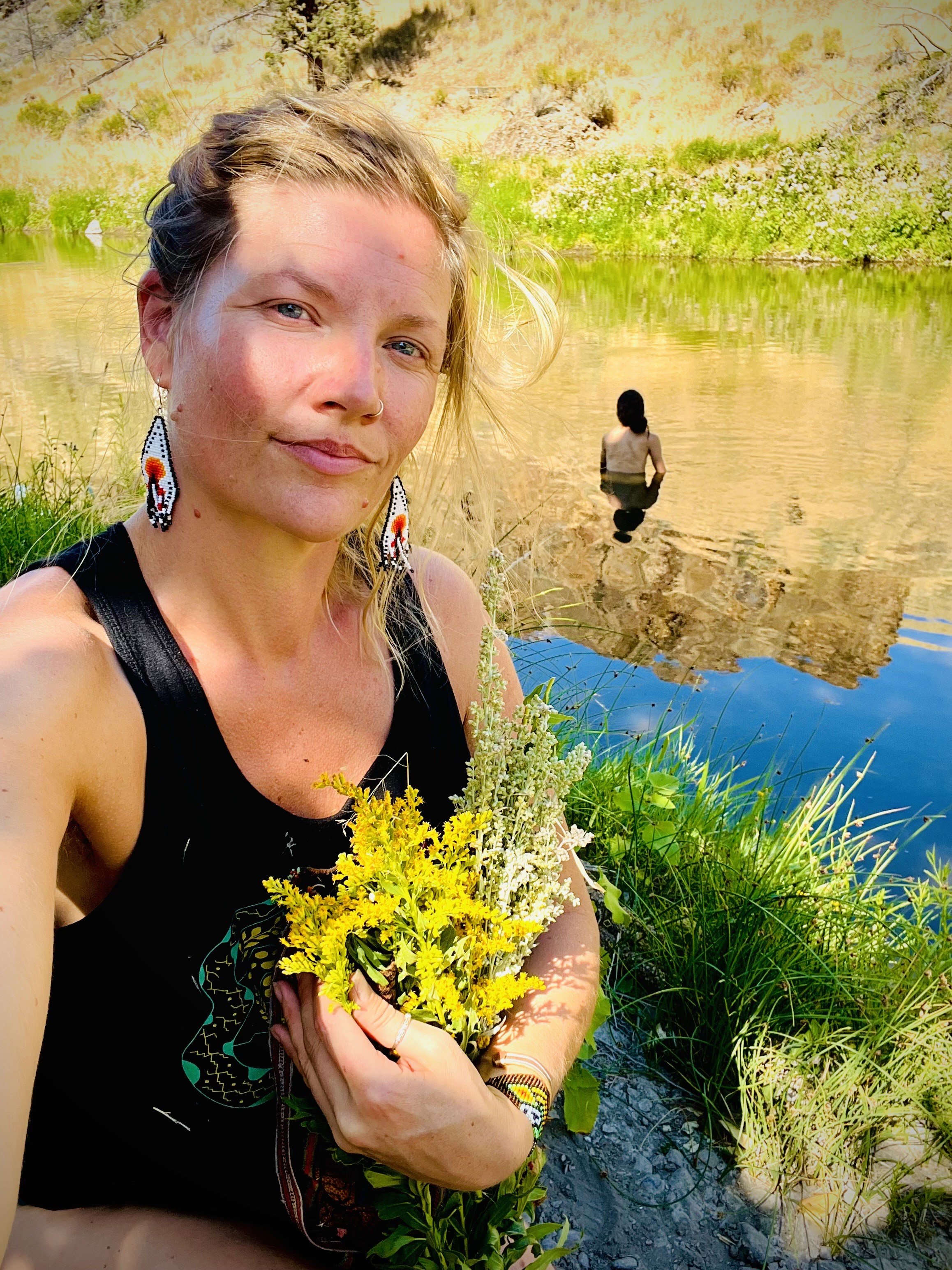 A woman taking a selfie by a river, holding a bouquet of wildflowers, with a person swimming in the water and a mountain in the background.