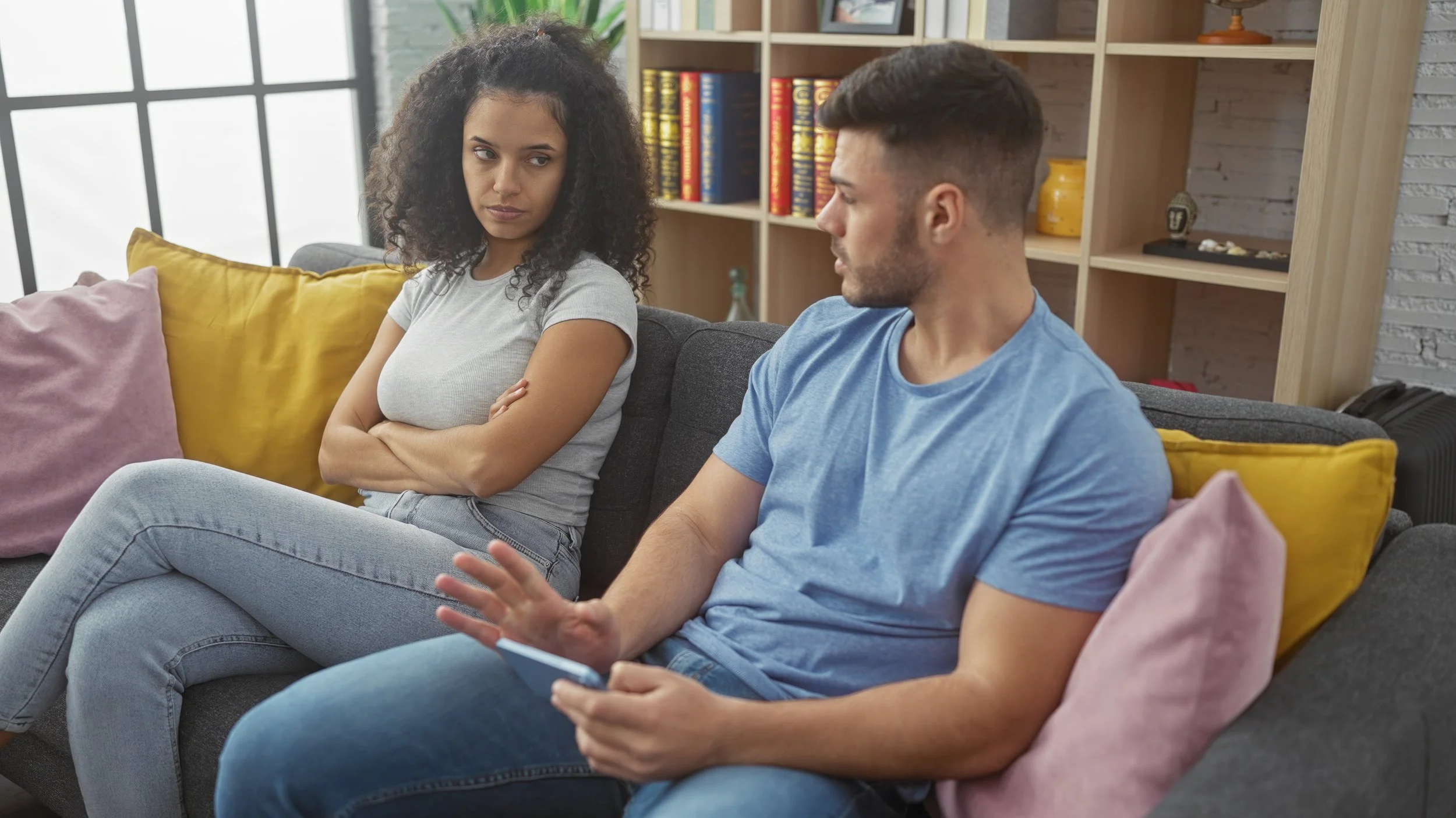 A couple sits on a couch in a living room. The woman has her arms crossed and looks upset while the man looks toward her and gestures with one hand, holding a phone in the other.