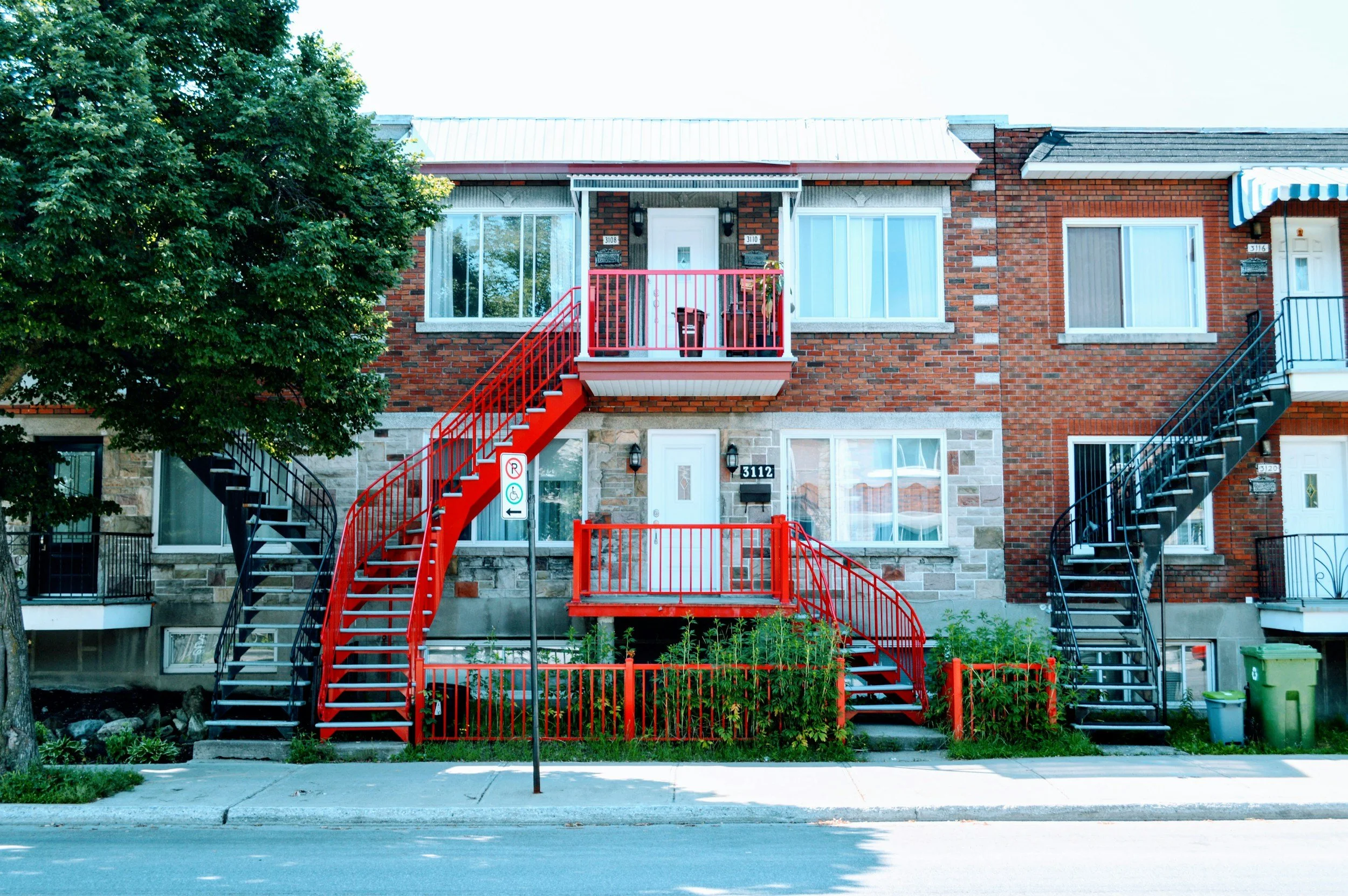 A multi-story brick apartment building with external stairs painted in red and black leading to small balconies. There are large windows, trees, a sidewalk, and a street in front.