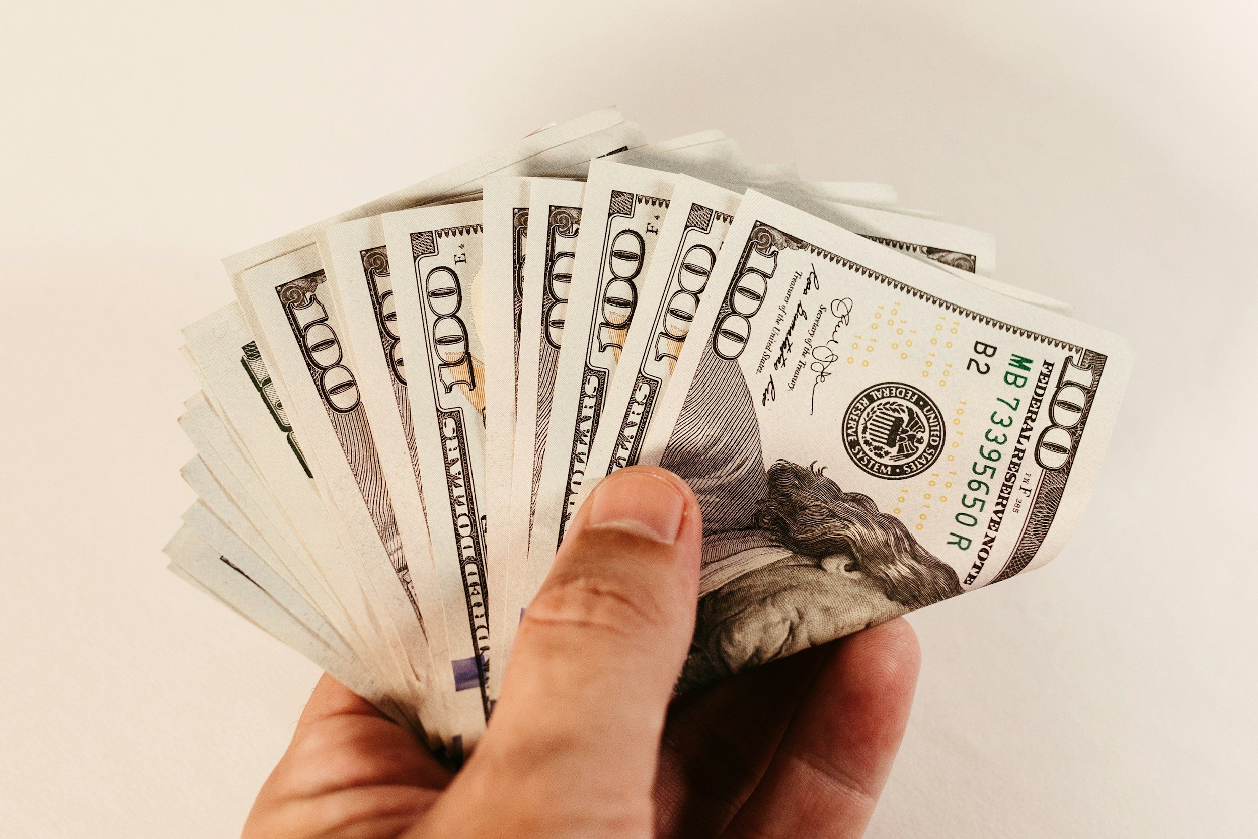 Hand holding a fan of ten $100 bills against a plain background.