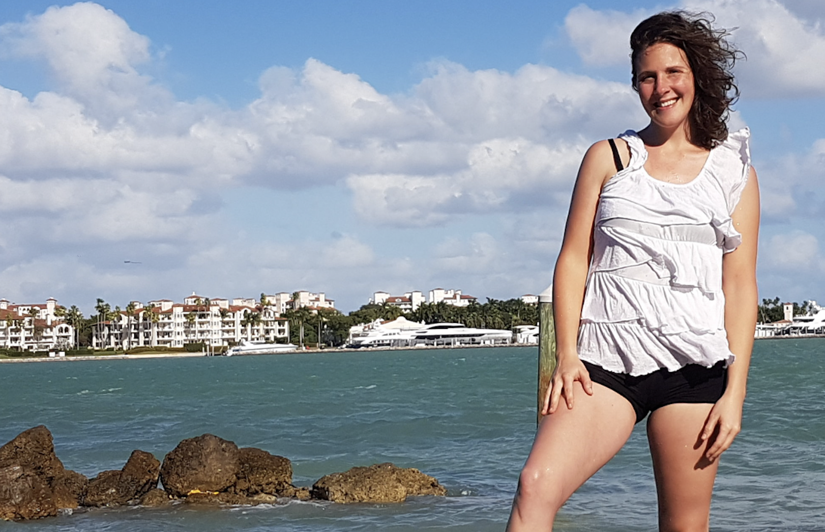 A woman standing on rocks by the water, with a city skyline and residential buildings in the background under a partly cloudy sky.