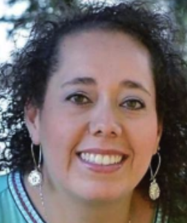 A close-up of a woman with curly dark hair, smiling, wearing earrings and a patterned top, outdoors with trees in the background.