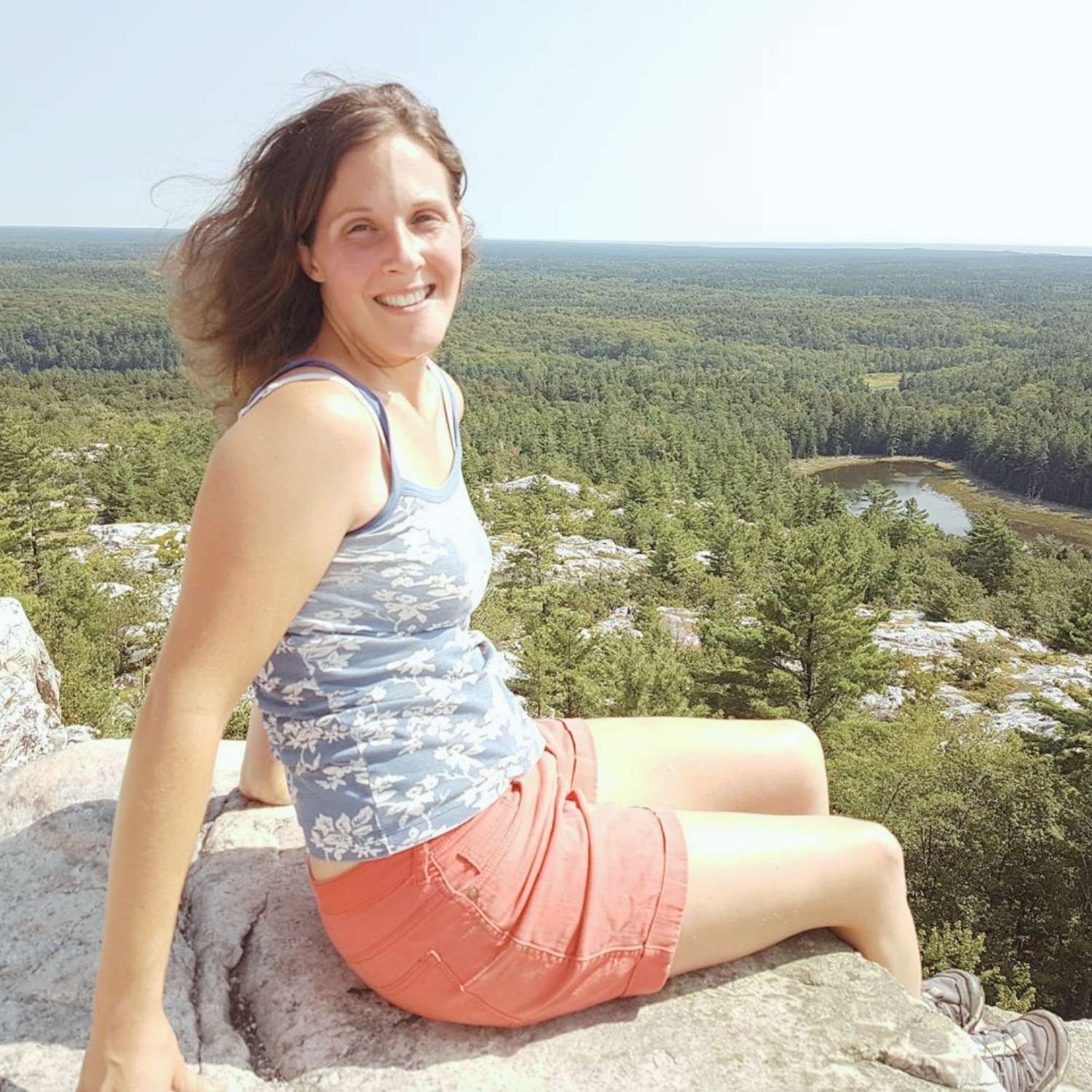Woman sitting on a rock overlooking a vast green landscape with trees, a pond, and a clear sky.