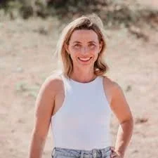 A woman smiling outdoors, wearing a white sleeveless top and light-colored jeans in a sandy area.