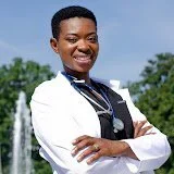 Portrait of a smiling woman in a white coat with arms crossed, outdoors with a fountain and trees in the background.