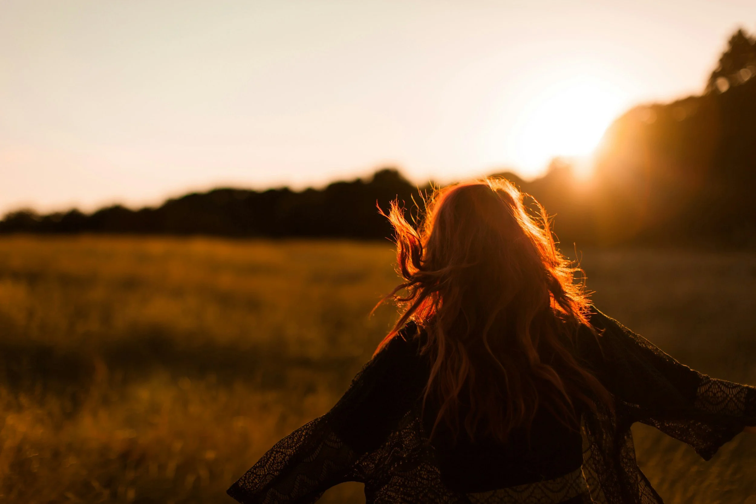 A person with long wavy hair sitting outdoors in a field at sunset, with arms outstretched, wearing a lace top.