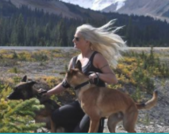 A woman with long white hair running in a field with three dogs, mountains and trees in the background.