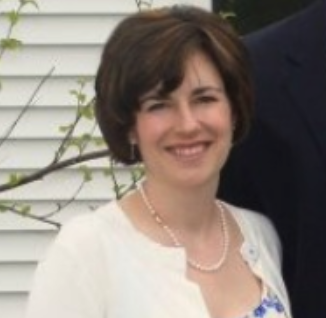 Smiling woman with short brown hair, wearing a white cardigan, pearl necklace, and floral dress, outdoors in front of a white fence.