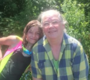 A young girl and an older woman standing outdoors with green foliage in the background, smiling at the camera.