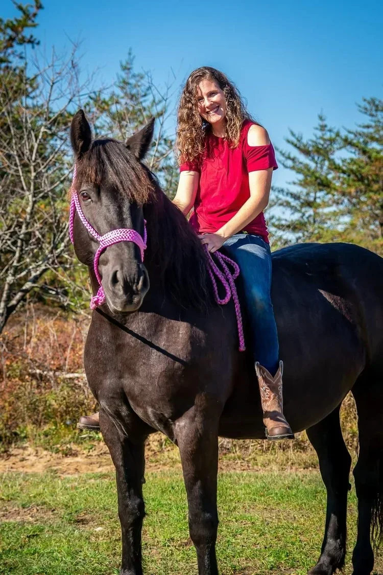 Sandie Gascon with curly hair wearing a red shirt and jeans riding a black horse with purple bridle and reins in an outdoor setting with trees and blue sky.