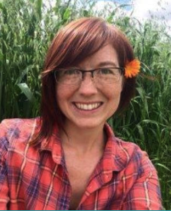 Woman with short reddish hair, glasses, and a flower in her hair, smiling outdoors in a field of tall green plants.