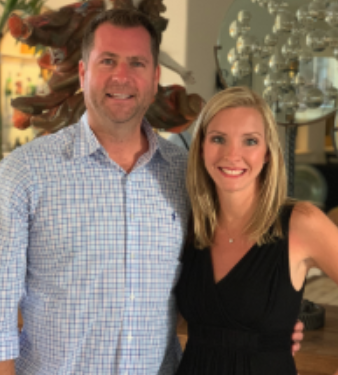 A man and a woman smiling and posing together indoors, with decorative items and artwork in the background.