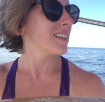A woman with short blonde hair wearing sunglasses and a purple tank top, smiling on a boat with water and sky in the background.