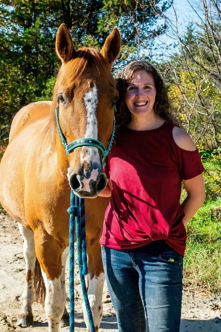 A woman standing next to a brown and white horse outdoors in a wooded area, smiling at the camera on a sunny day.