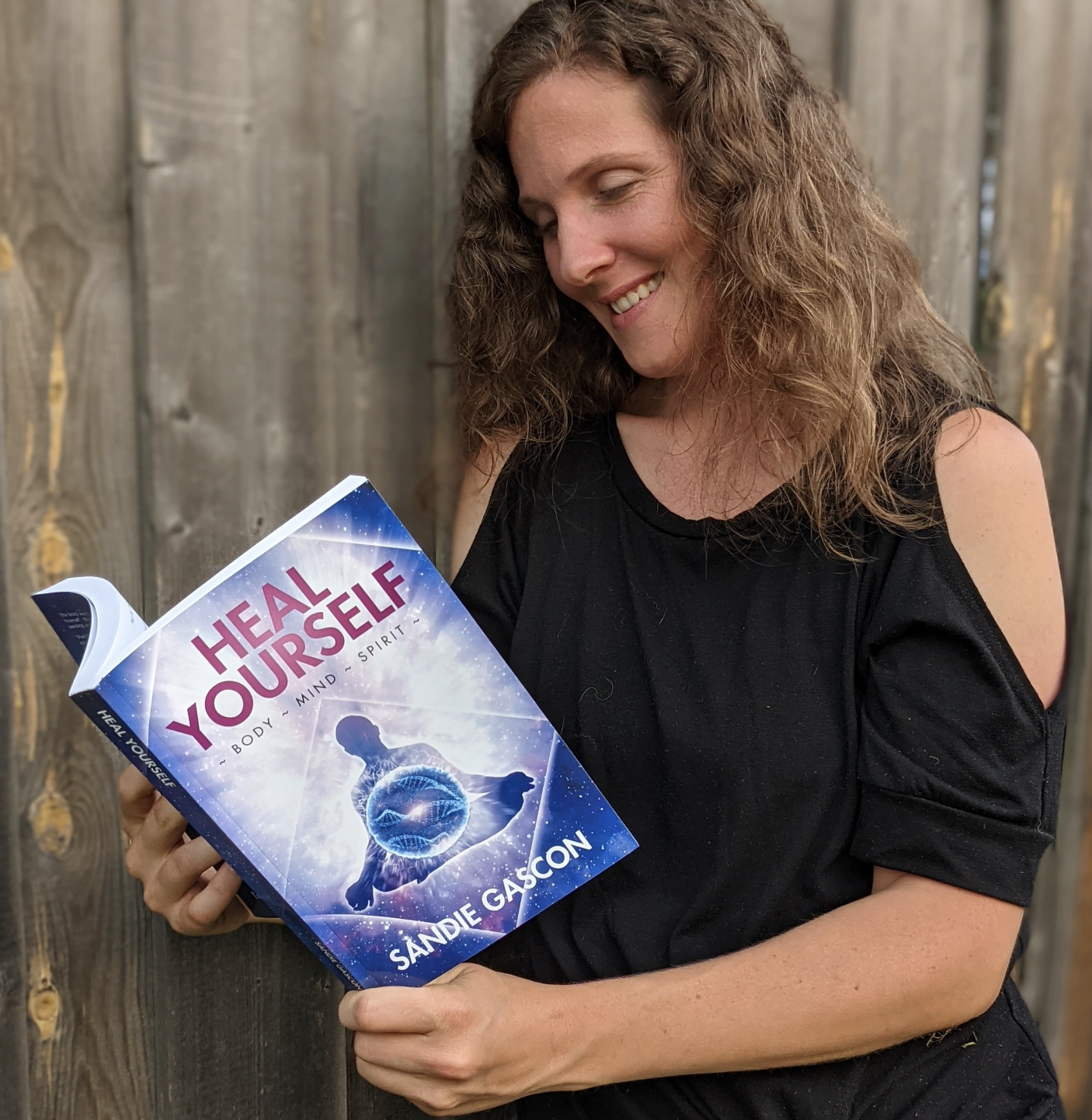 A woman with curly hair, wearing a black top, smiling while looking at a book titled 'Heal Yourself' by Sandie Gascon, with a light-colored wooden fence in the background.
