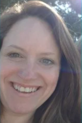 Close-up of a smiling woman with light-colored eyes and long brown hair outdoors on a sunny day.