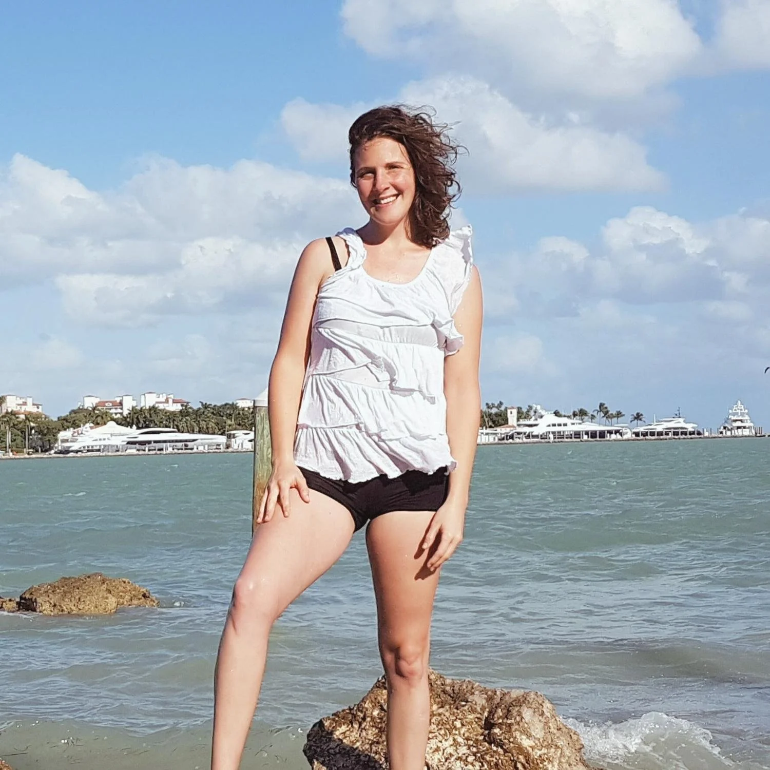 A woman standing on rocks at the beach, smiling, with water and buildings in the background, under a partly cloudy sky.