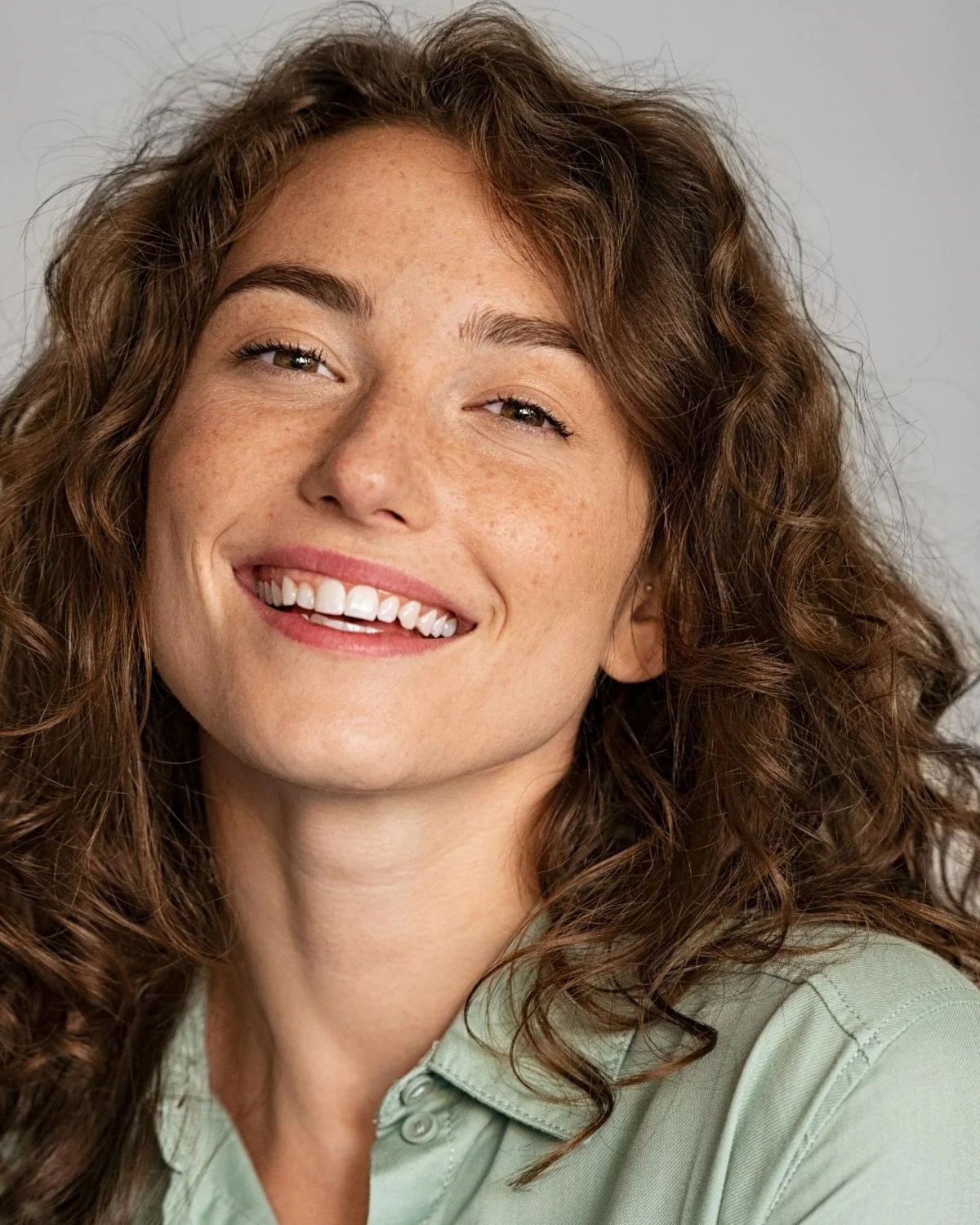 Close-up portrait of a smiling woman with wavy brown hair, freckles, and light skin, wearing a light green shirt, against a plain gray background.