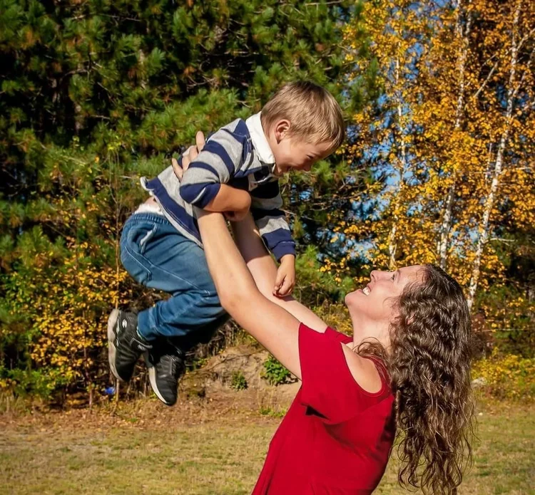 A woman in a red shirt lifts a young boy into the air outdoors, both smiling, with colorful fall trees in the background.