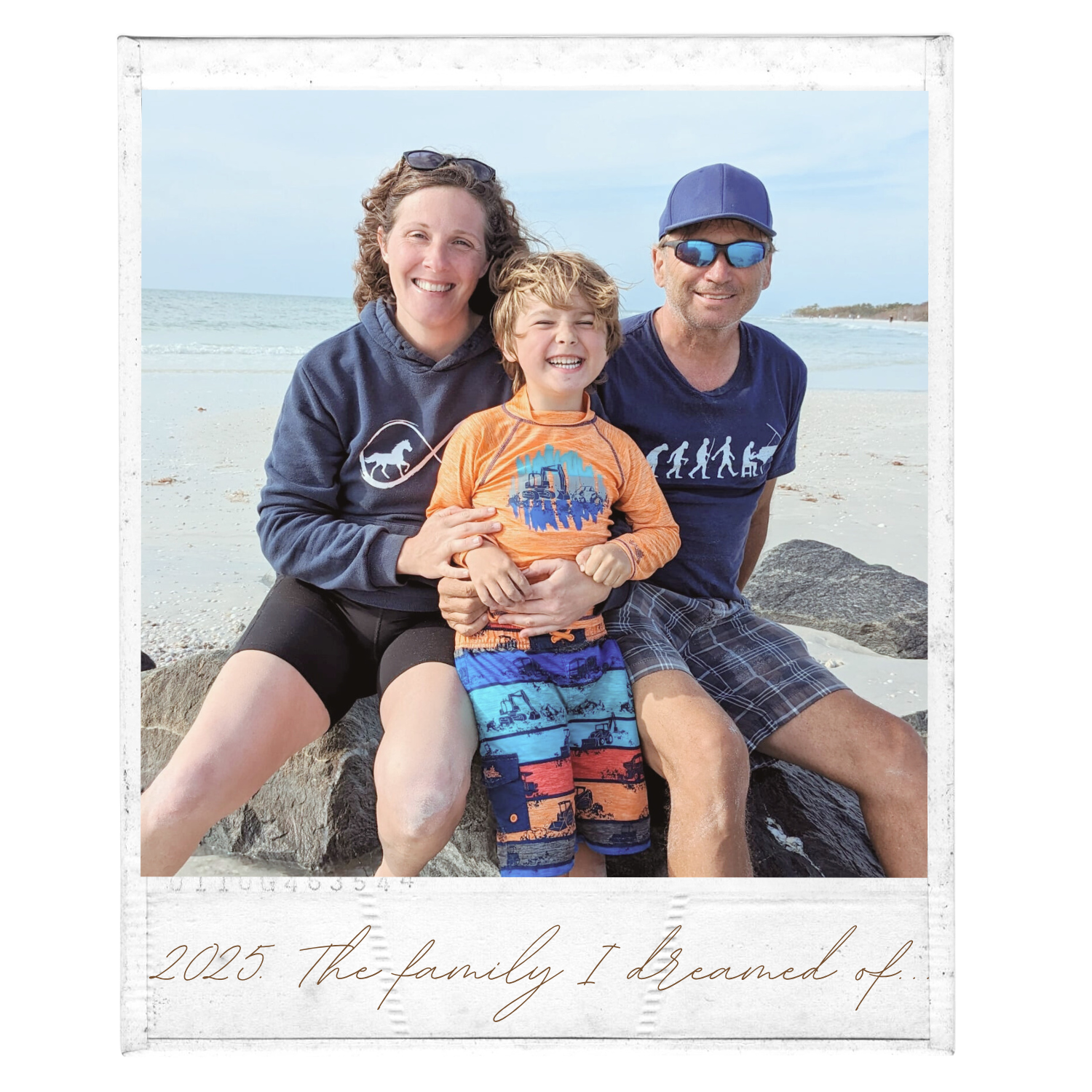 A family of three sitting on rocks by the beach, smiling. The woman is on the left, wearing a navy hoodie. The young boy is in the middle, wearing an orange shirt with a cityscape and colorful shorts. The man is on the right, wearing a blue cap, sunglasses, a blue T-shirt, and plaid shorts. The background features the ocean, sandy beach, and a cloudy sky. The image has a caption at the bottom reading "2025. The family I dreamed of."