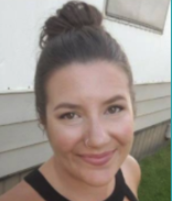 Close-up of a young woman with brown hair tied in a bun, smiling at the camera, standing outdoors near a white building.