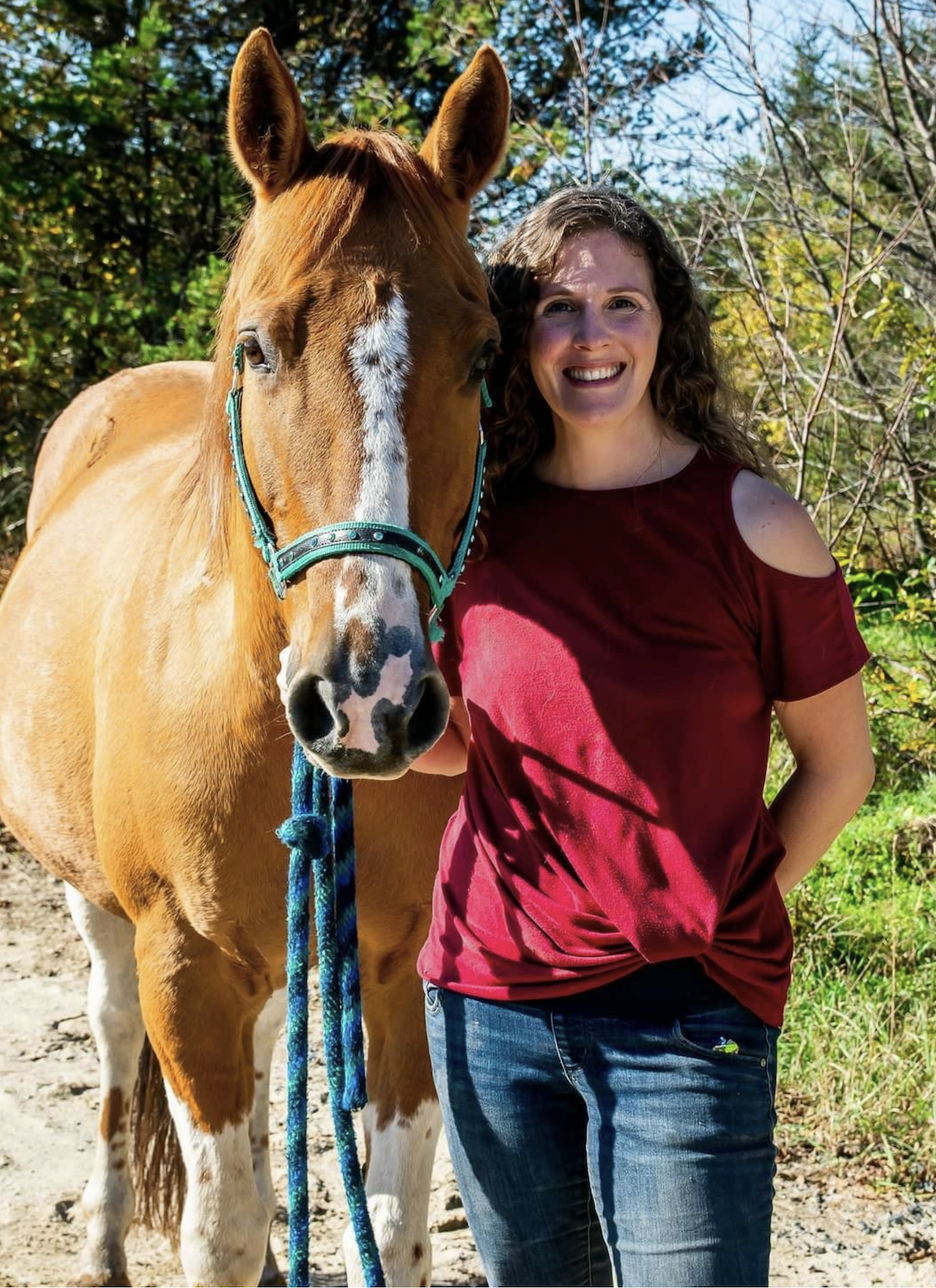 A woman with curly brown hair wearing a red cold-shoulder top and blue jeans standing beside a chestnut horse with a white face marking and blue halter in a sunny outdoor setting with green foliage.